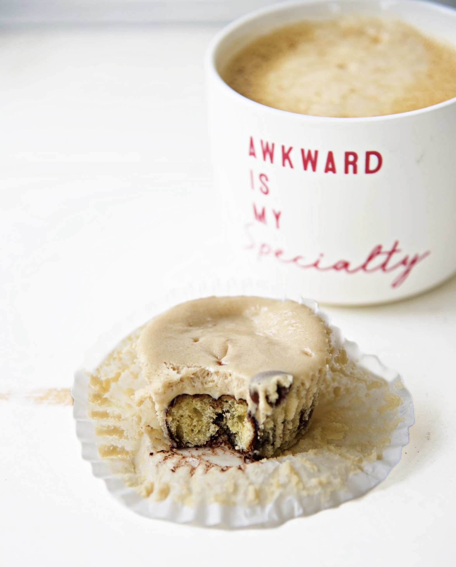 Coffee and Donuts Cheesecake Bite with the cupcake liner pulled back exposing half of the cheesecake bite. With a cup of coffee in the upper right hand corner with a cup that reads Awkward is my specialty. 