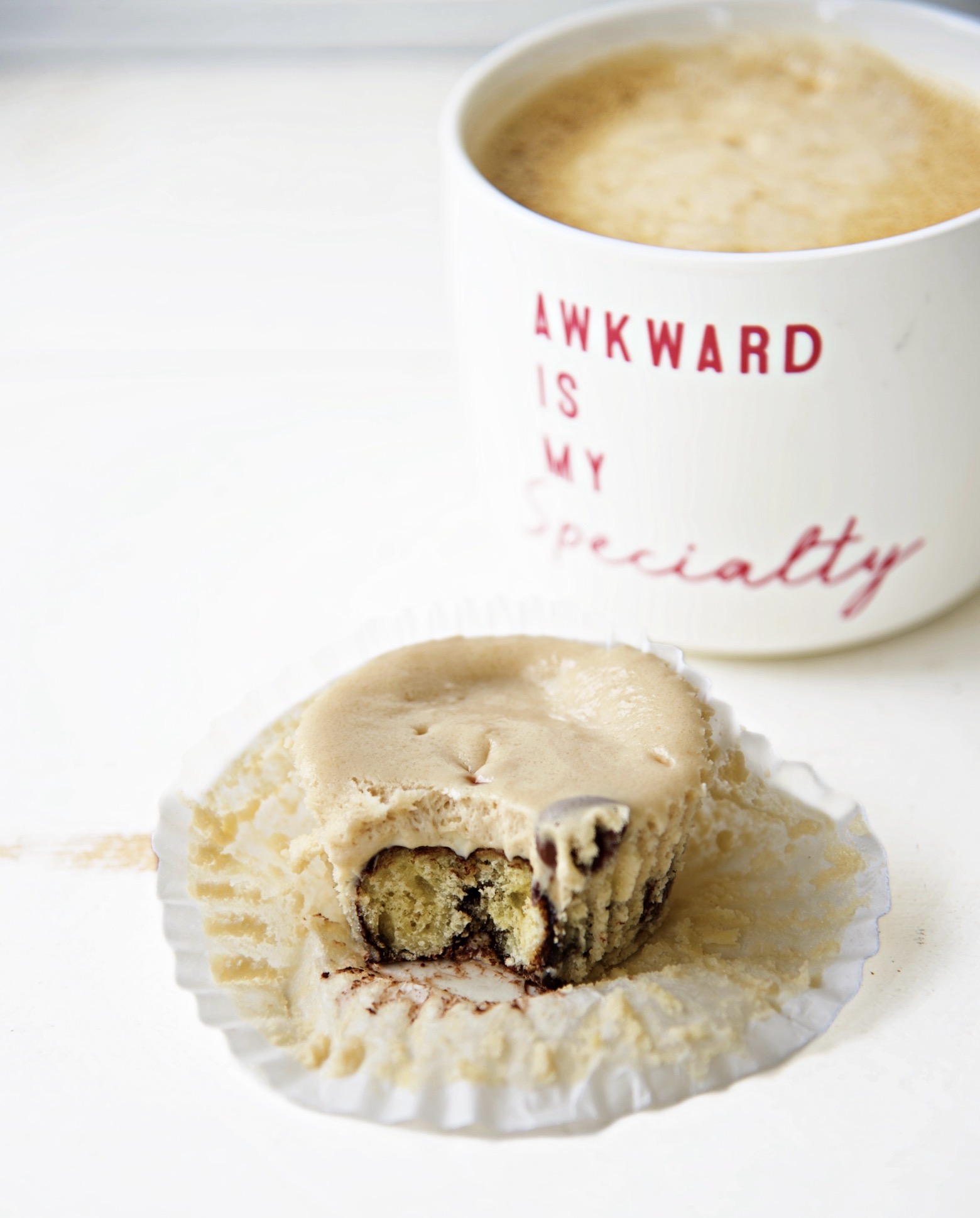 Coffee and Donuts Cheesecake Bite with the cupcake liner pulled back exposing half of the cheesecake bite. With a cup of coffee in the upper right hand corner with a cup that reads Awkward is my specialty.