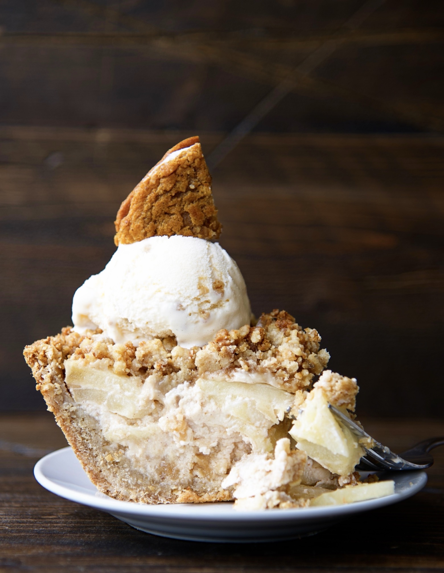 Side view of piece of the Oatmeal Creme Apple Pie on a small white plate with ice cream and a cookie pie wedge on top of the pie with a fork having taken a piece of the pie lays on the plate. 