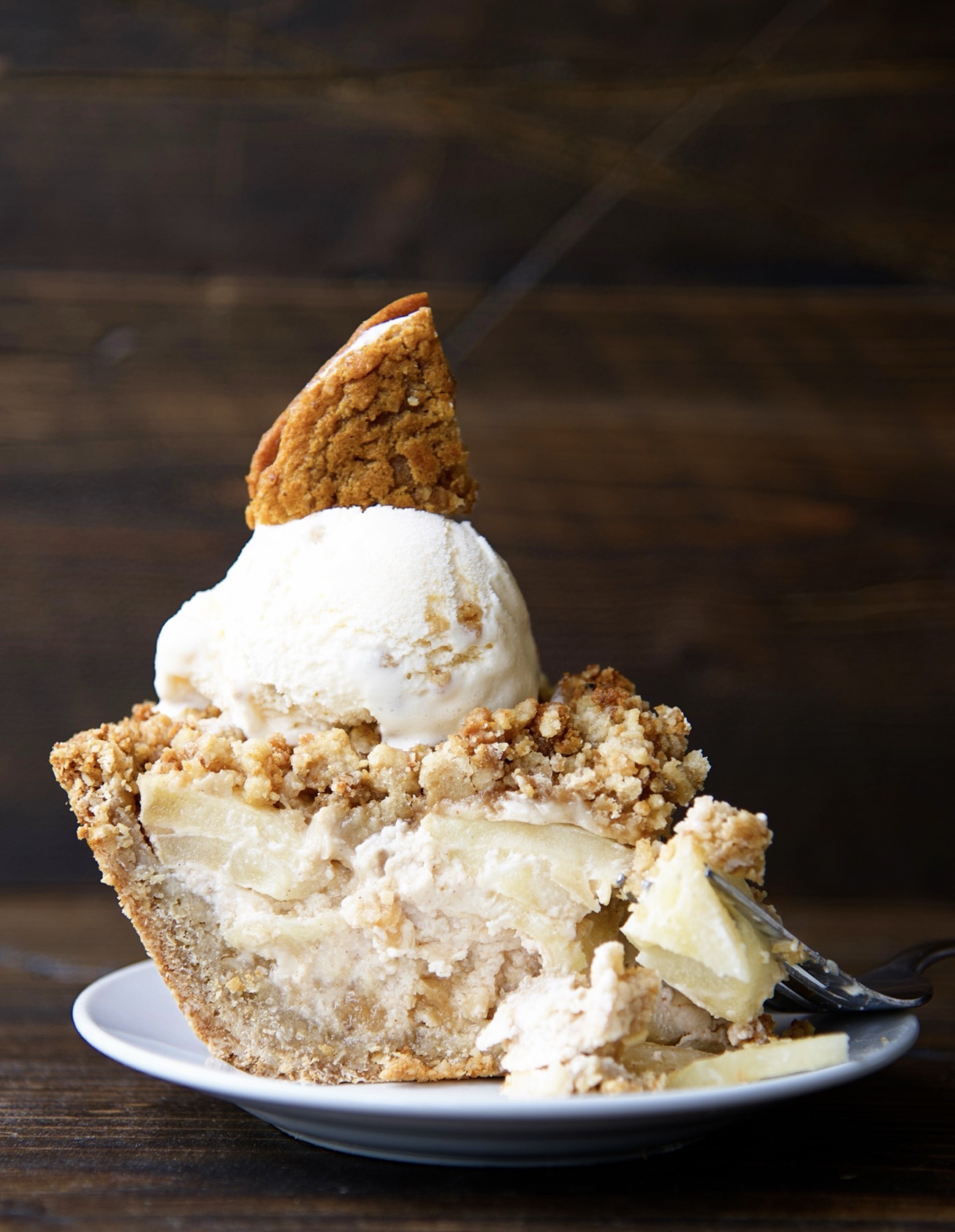 Side view of piece of the Oatmeal Creme Apple Pie on a small white plate with ice cream and a cookie pie wedge on top of the pie with a fork having taken a piece of the pie lays on the plate.