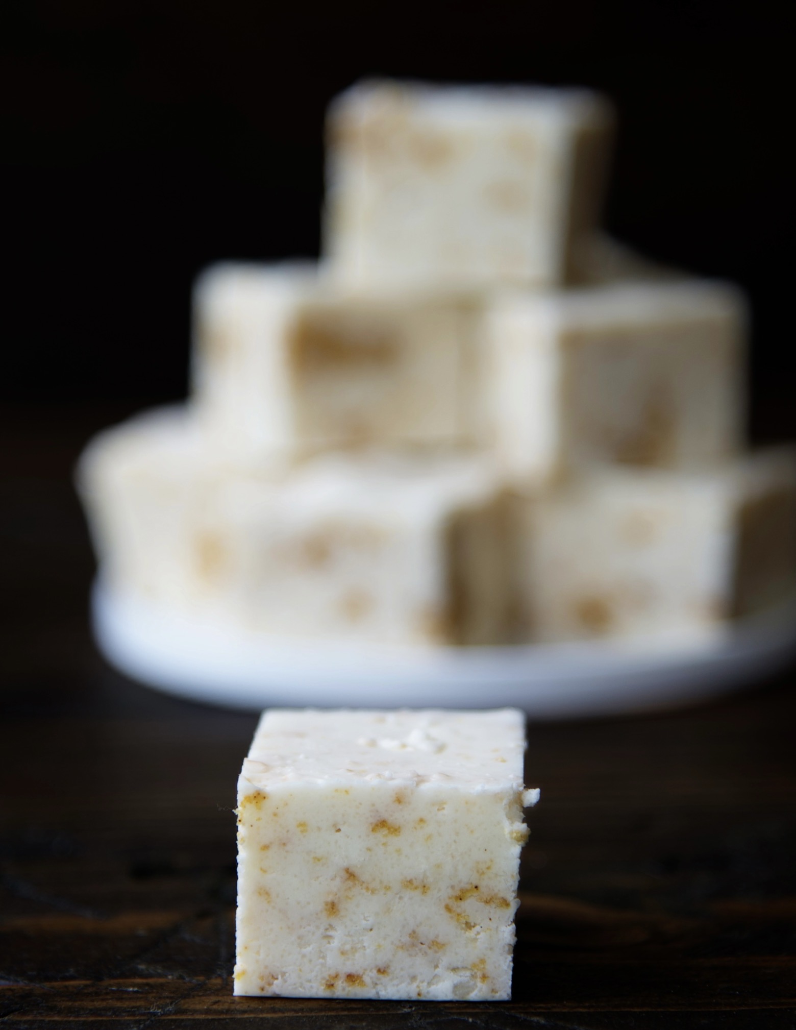 Single piece of fudge up front with a small plate of fudge blurred in the background. 