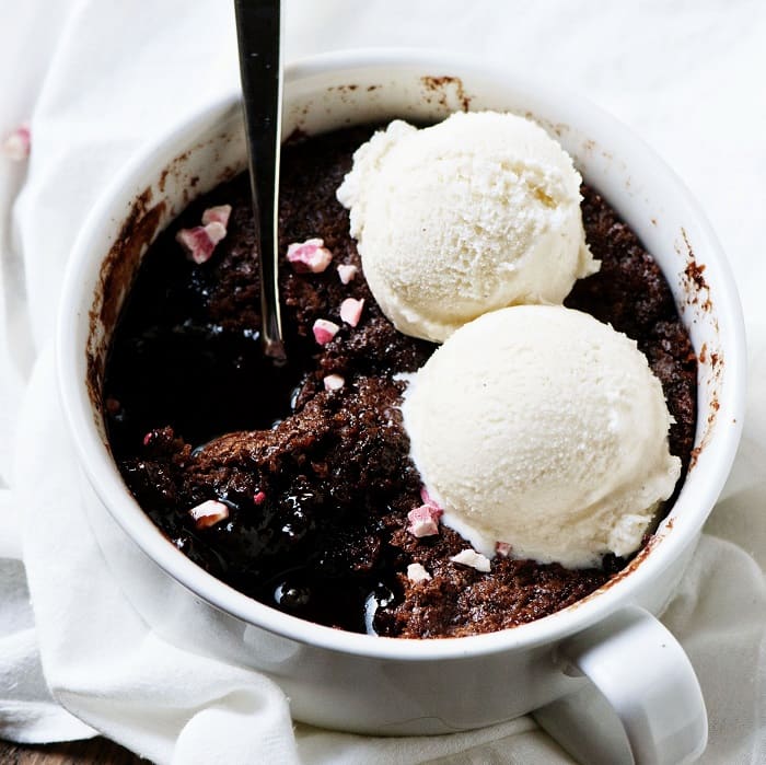 Close up of the cobbler with sprinkled peppermint crunch chips on top with two scoops of ice cream with a spoon in it exposing the chocolate sauce below the cobbler. 