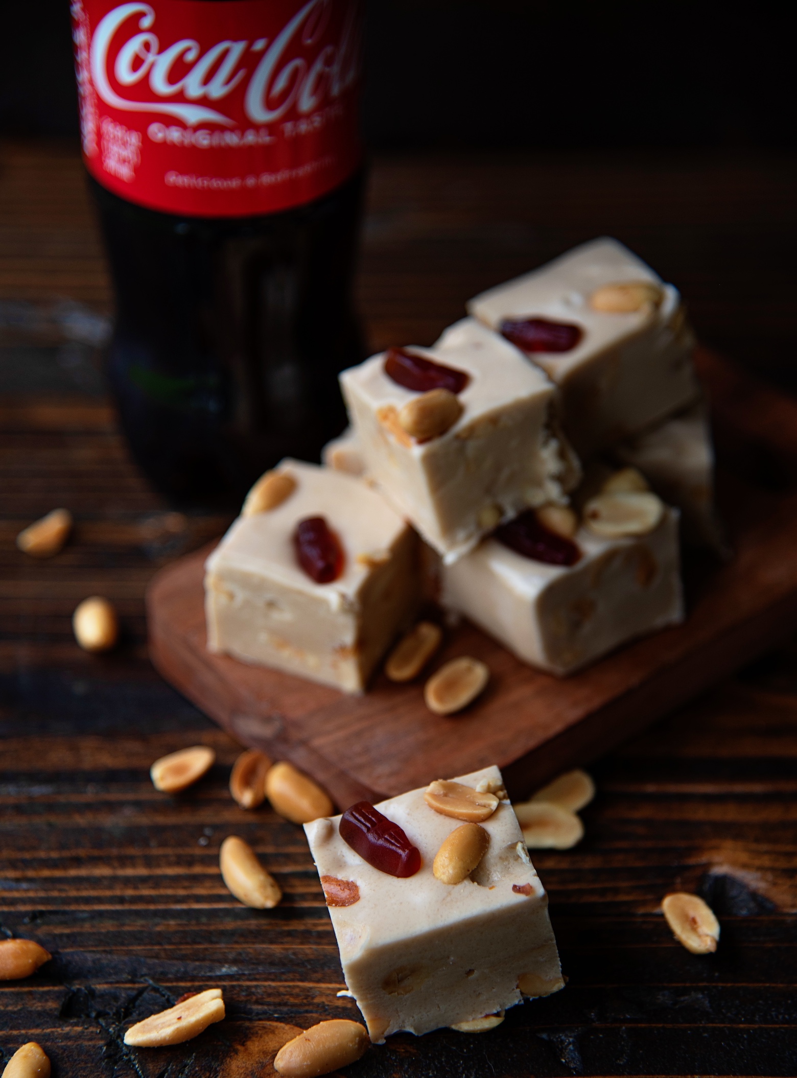 Pieces of Coke and Peanut Fudge on a small cutting board with one piece of fudge in front of it and a bottle of Coke behind the fudge. 