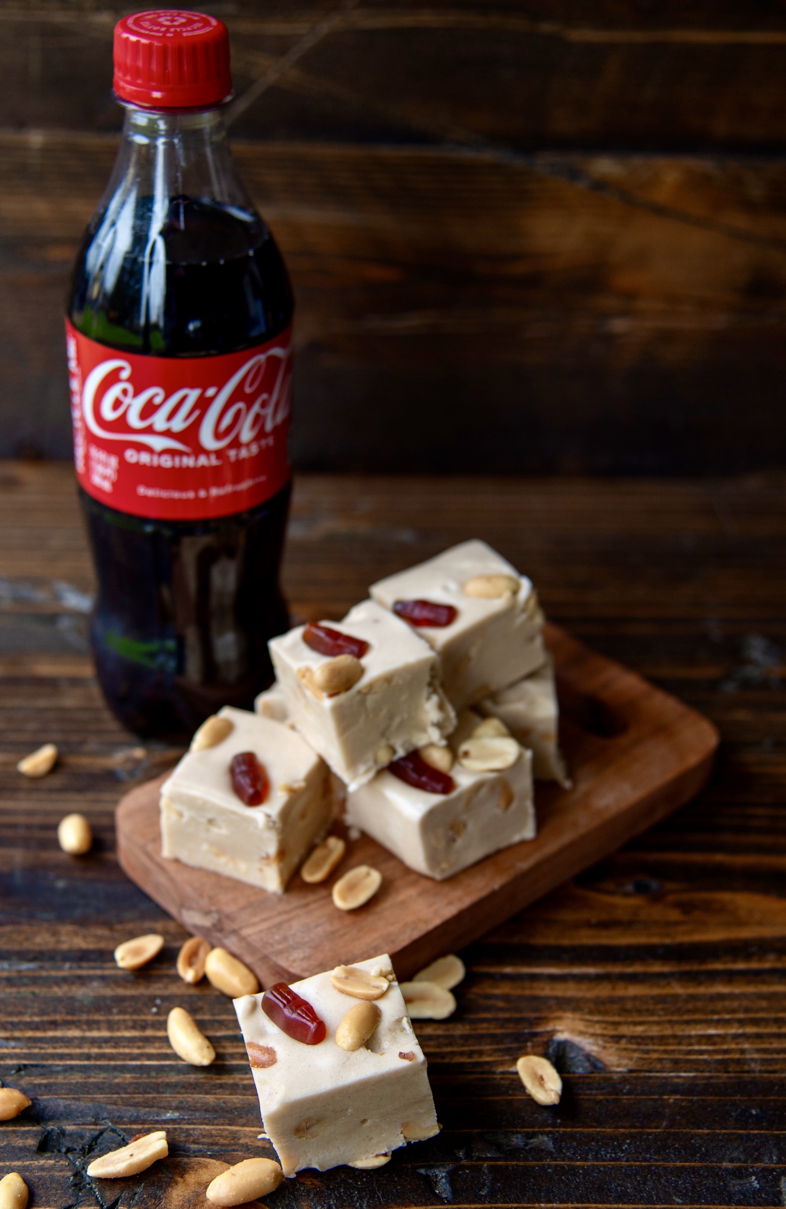 Pieces of Coke and Peanut Fudge on a small cutting board with one piece of fudge in front of it and a bottle of Coke behind the fudge. 