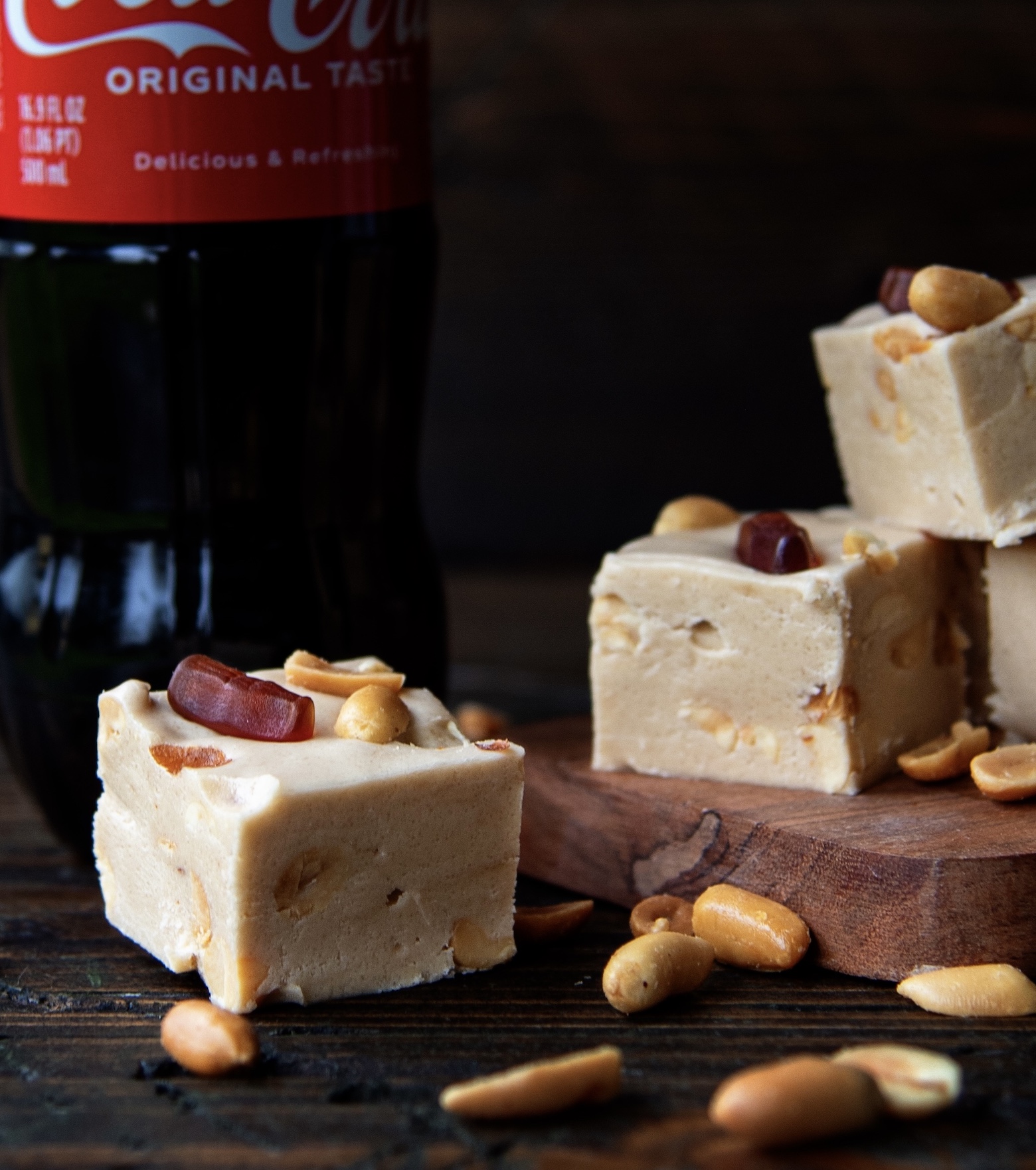 Pieces of Coke and Peanut Fudge on a small cutting board with one piece of fudge in front of it and a bottle of Coke behind the fudge. 