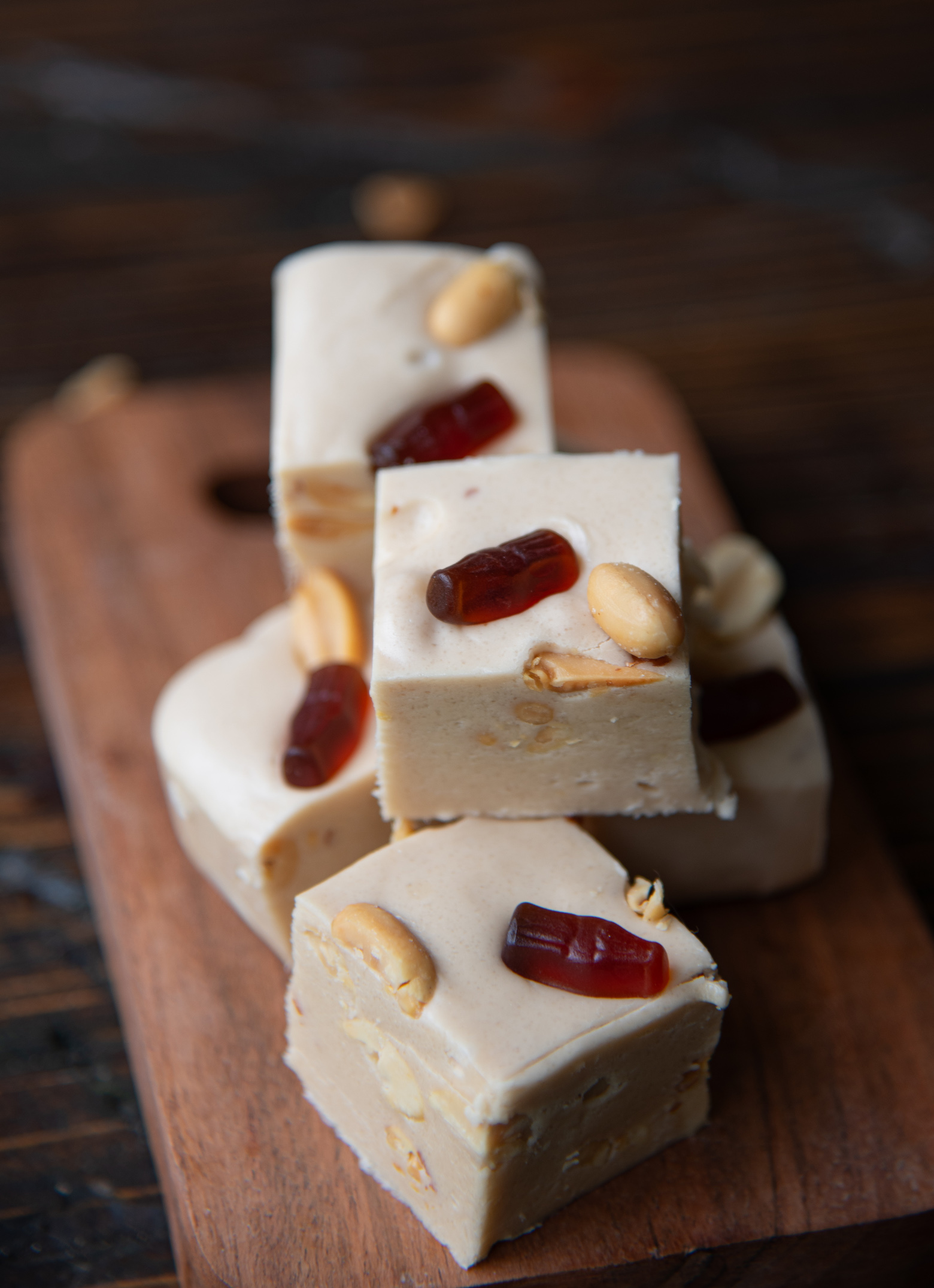 Pieces of fudge stacked on a small wooden cutting board. Focus on the peanuts and mini gummy cola on top of the fudge.