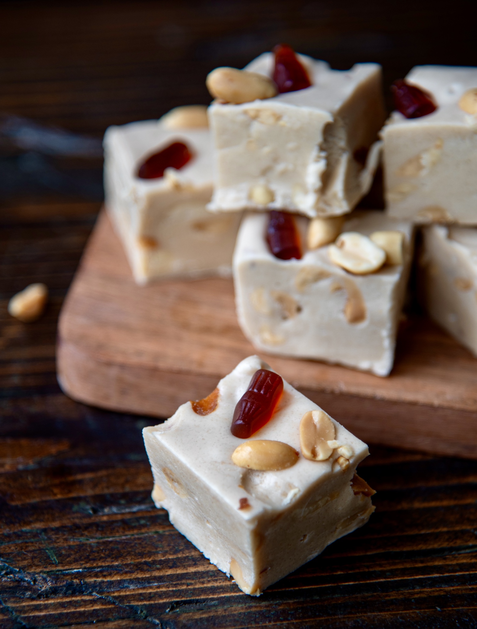 Close up of a piece of fudge with other pieces of fudge stacked on a small wooden cutting board. Focus on the peanuts and mini gummy cola on top of the fudge.