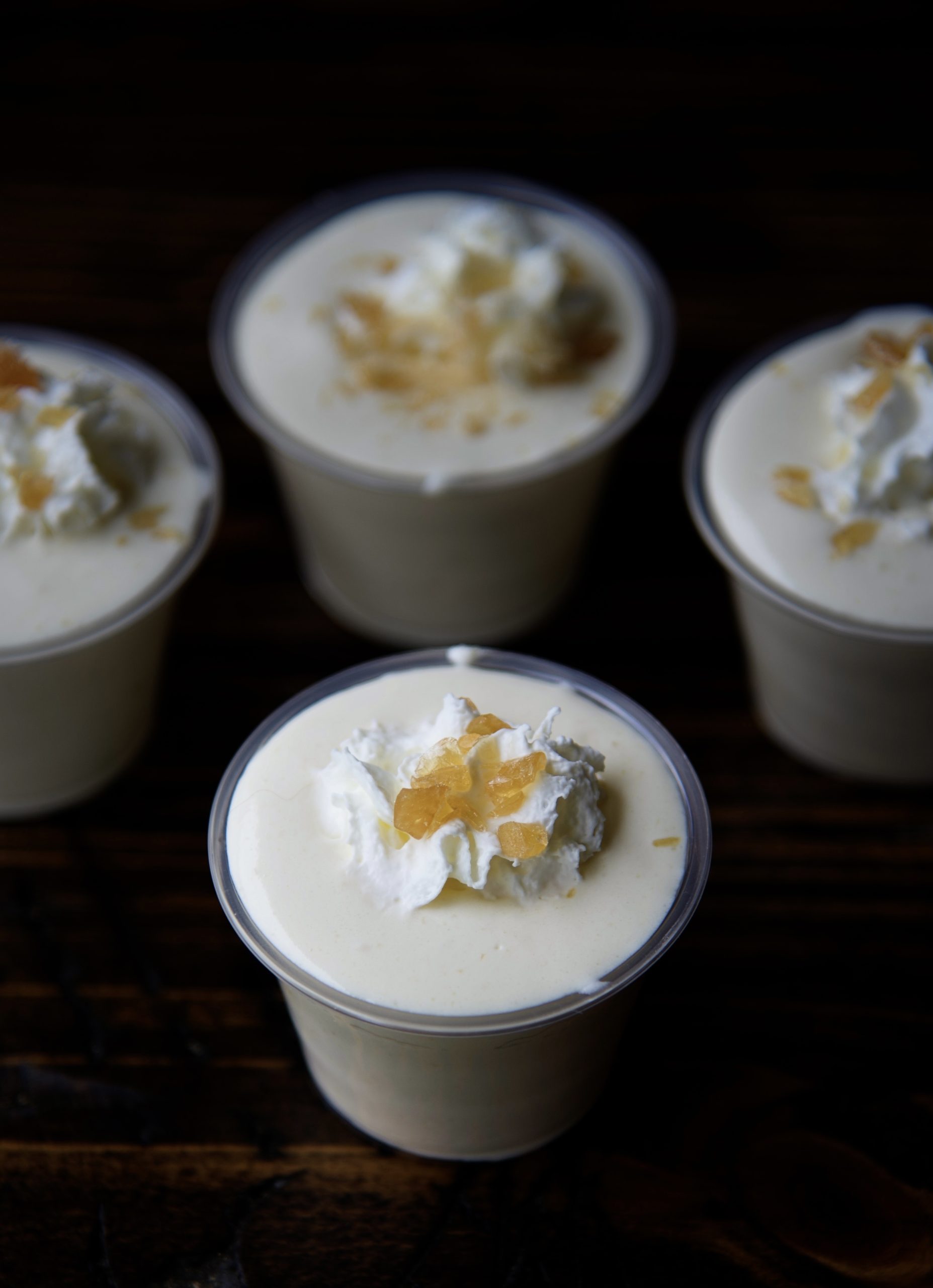 Four pudding shots with the center one being the focus. Shots are topped with whipped cream and crushed butter rum candies. 