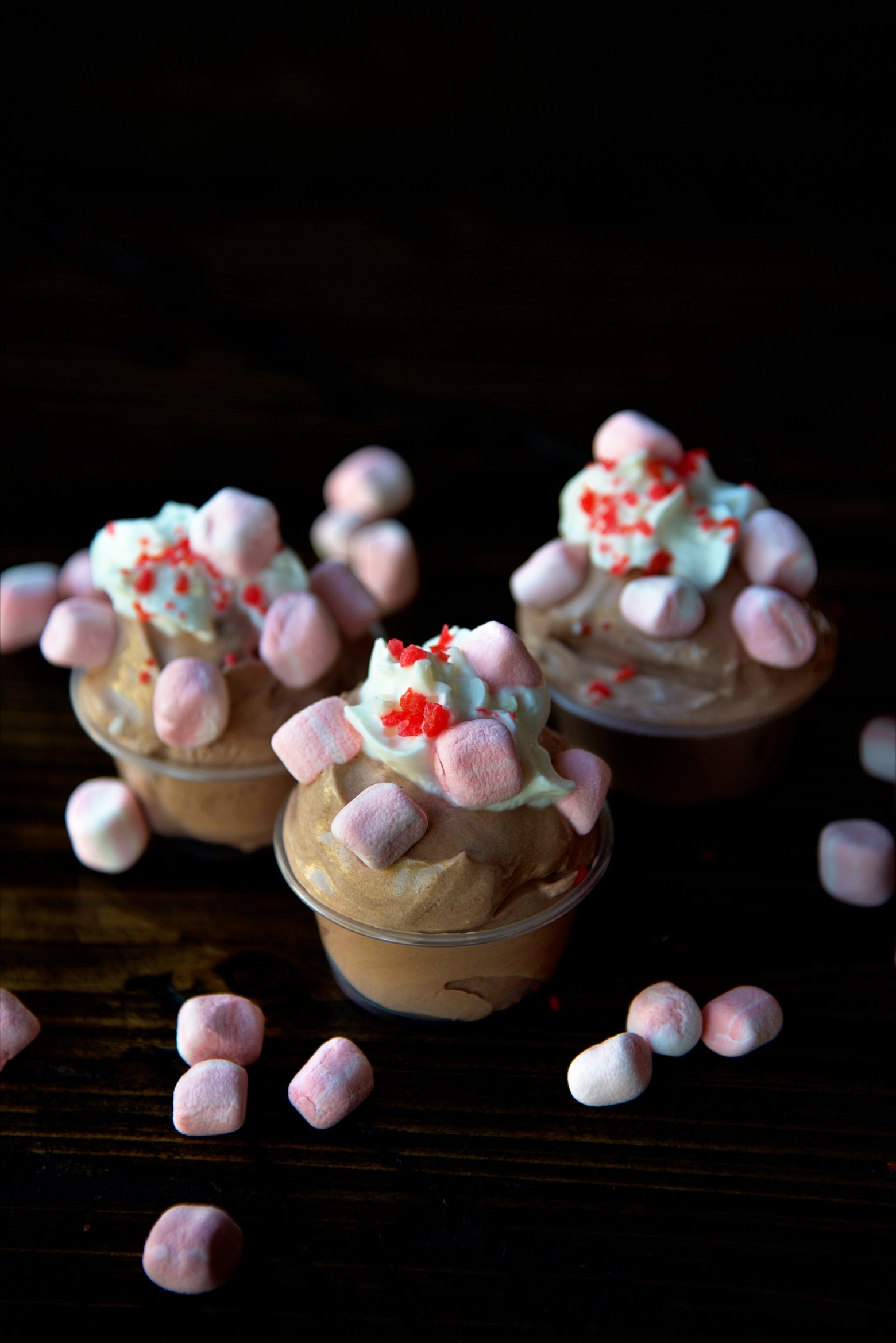Three quarter view of three pudding shots with whipped cream. peppermint marshmallows, and pop rocks on each one. Peppermint marshmallows scattered around the shots. 