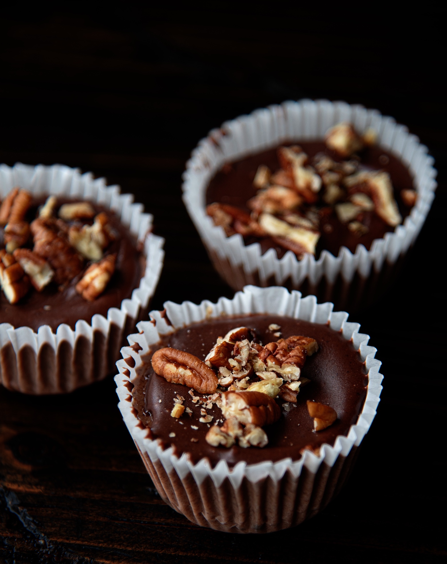 Three whole Cheesecake Bites with focus on the center cheesecake bite. Focus on the top of the cheesecake bites that have chocolate icing and pecans. 