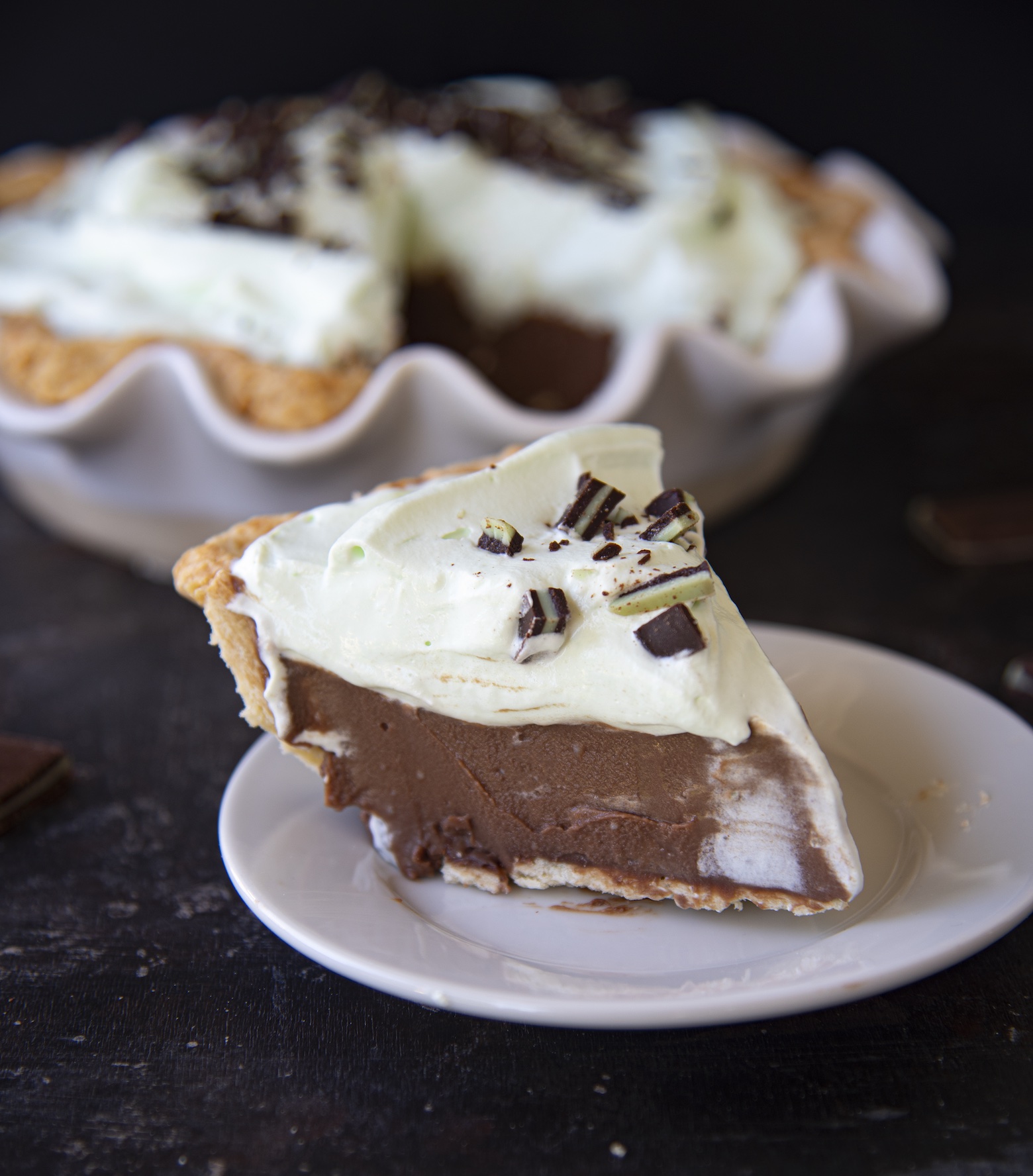 Slice of Andes Mint Chocolate Cream Pie on a small white plate out in front of the whole pie which is in the background. 