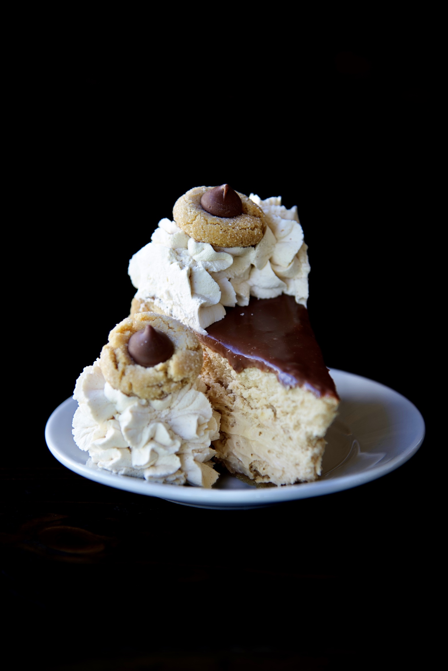 Single piece of cheesecake on a small white plate. With peanut butter whipped cream on the side and on top of the cheesecake. Topped with mini peanut blossom cookies. 