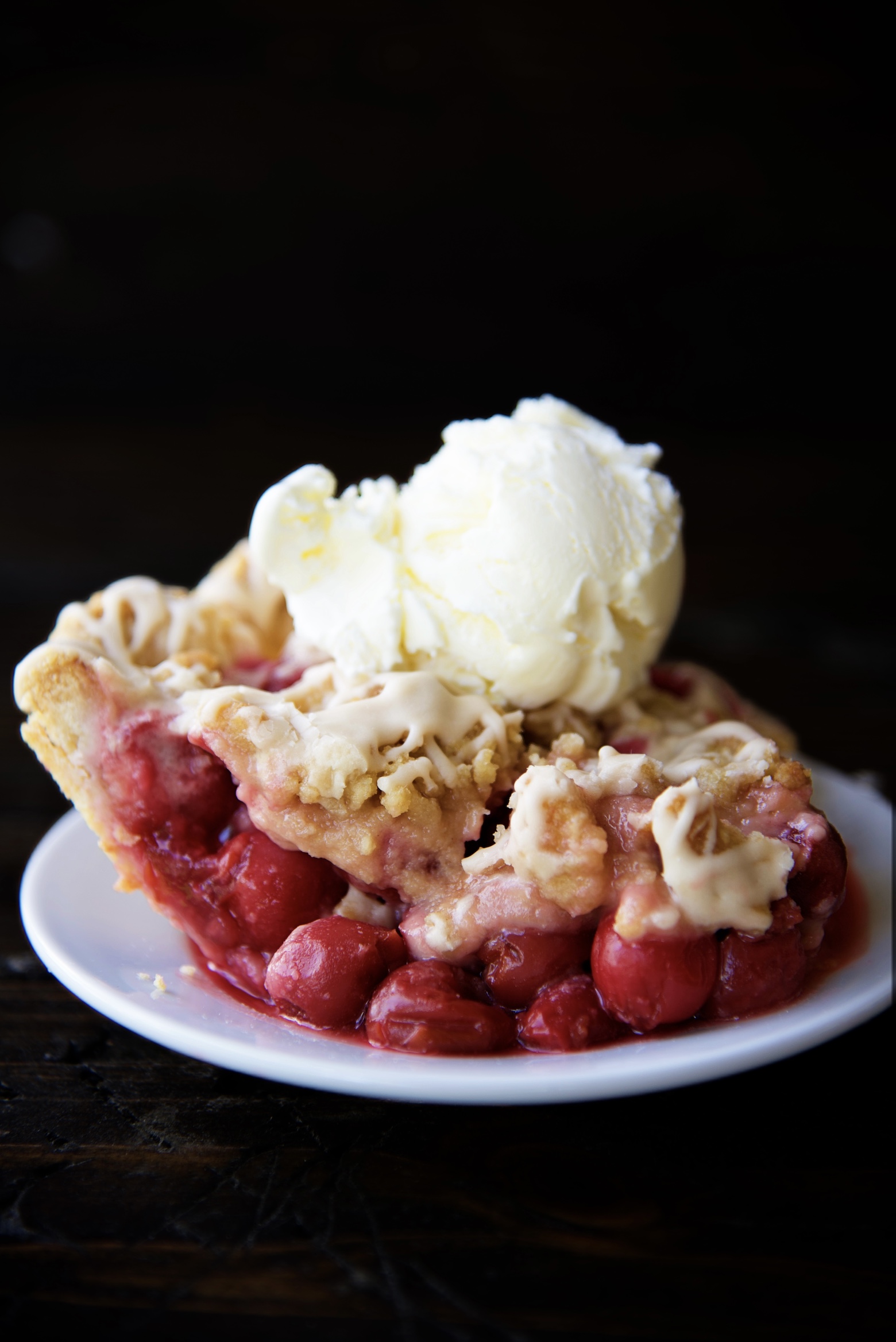 Close up of a slice of Cherry Cola Crumb Pie on a small white plate with a scoop of vanilla ice cream on top. 