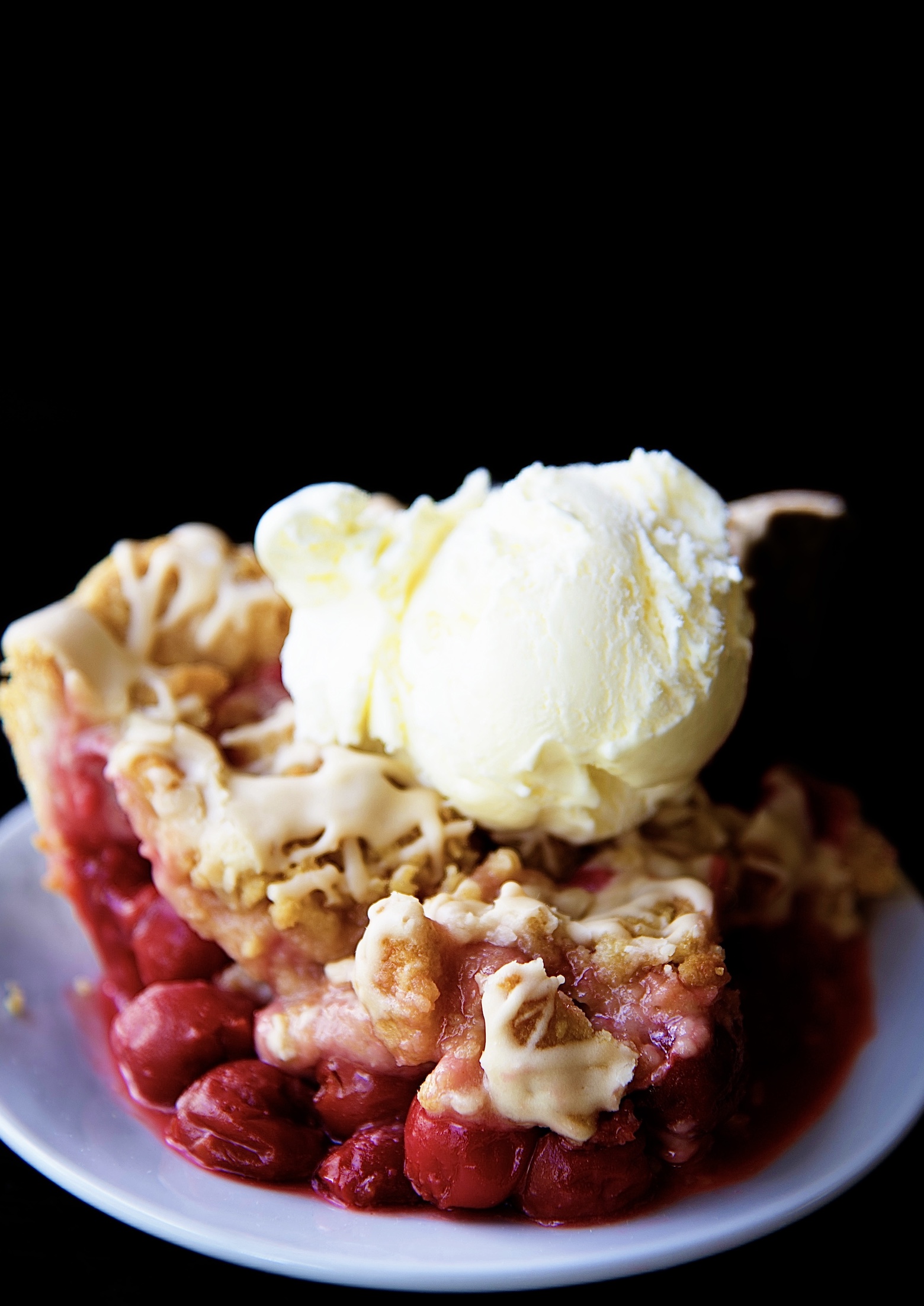Close up of a slice of Cherry Cola Crumb Pie on a small white plate with a scoop of vanilla ice cream on top. 