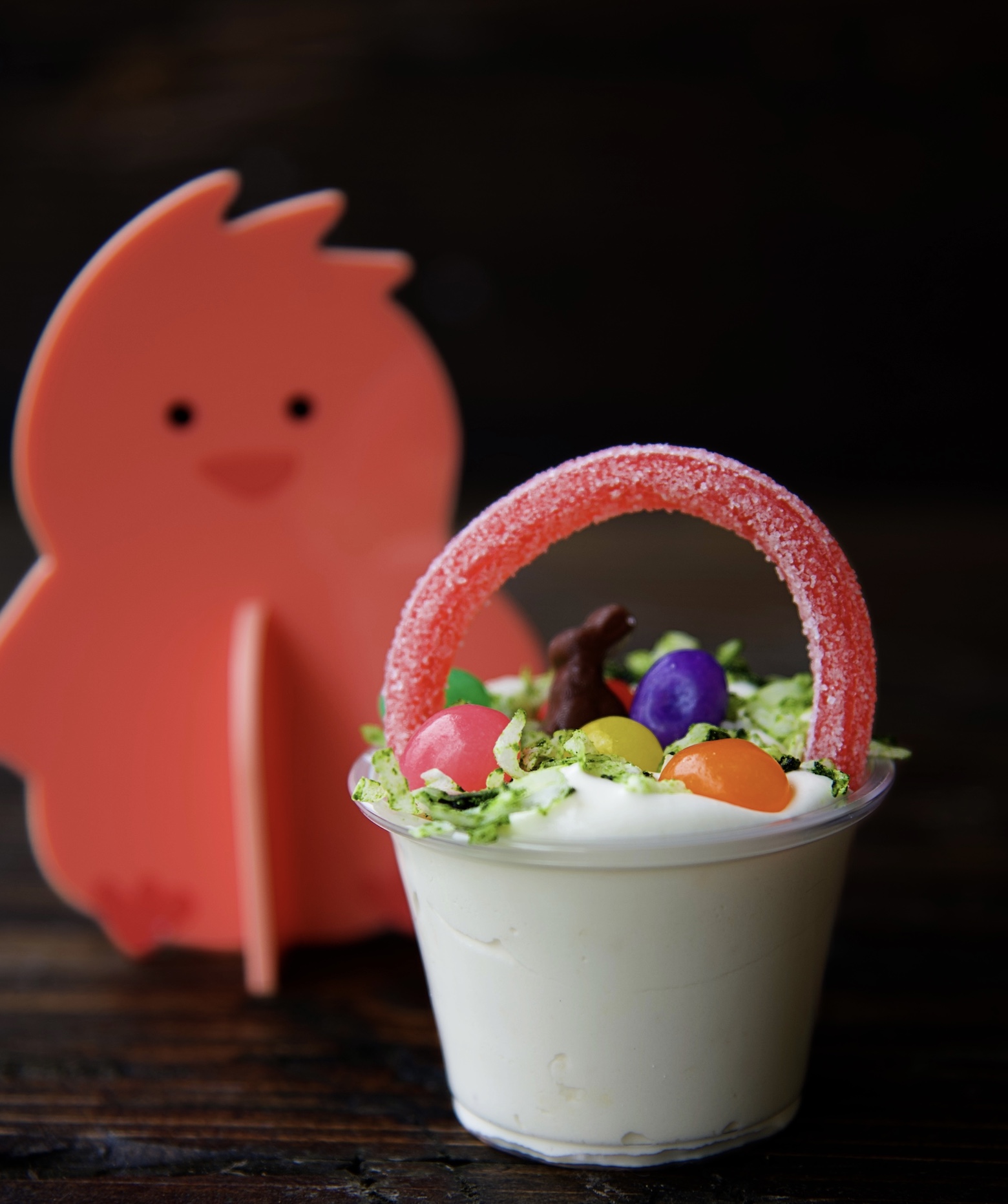 Side view of a single pudding shot with with licorice handle, coconut grass, mini chocolate bunny, and mini jelly beans. There is a pink chick in the background. 