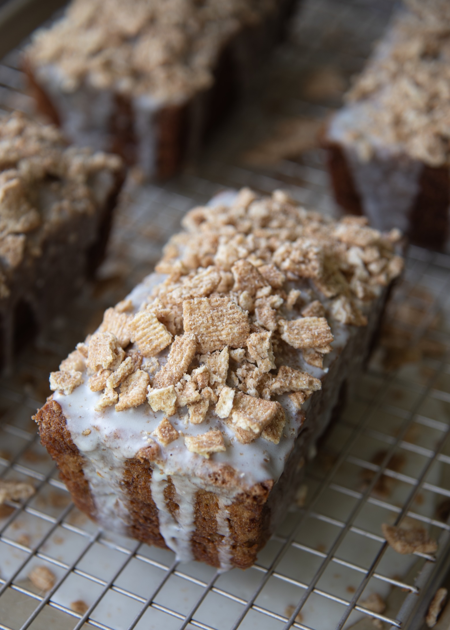 This photo features a close-up of a mini loaf of Cinnamon Toast Crunch Banana Bread resting on a wire cooling rack. The loaf is generously topped with a thick layer of cereal milk glaze that drips down the sides, creating a luscious, glossy finish. Piled high on top is a crunchy layer of crushed Cinnamon Toast Crunch cereal, adding texture and a nostalgic, cinnamon-sugar flair. In the background, other similarly topped mini loaves are slightly out of focus, giving the image a warm, inviting, and bakery-fresh feel. The lighting highlights the golden brown crust and the creamy glaze, making the loaf look irresistibly delicious.