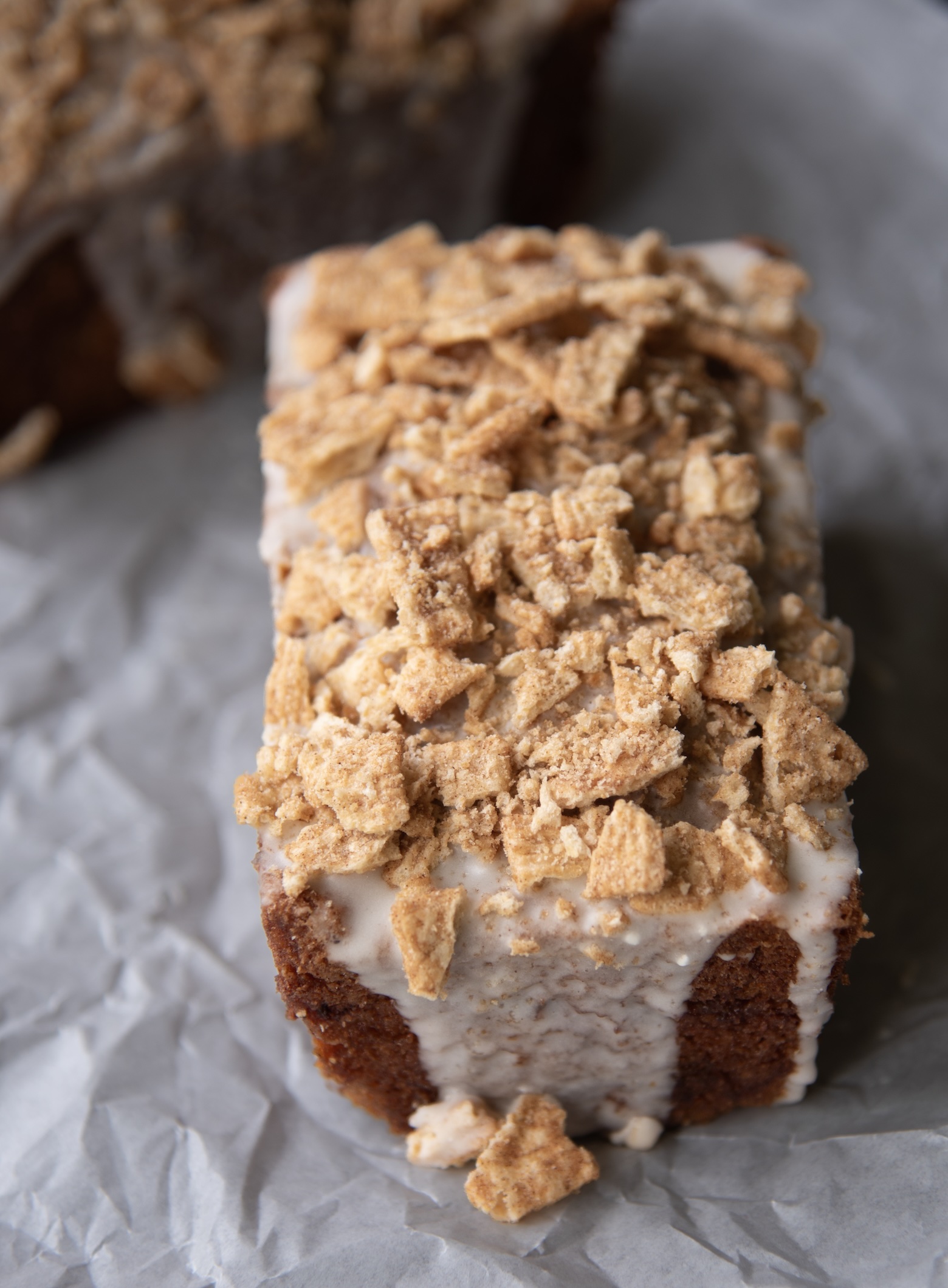 This photo features a single mini loaf of Cinnamon Toast Crunch Banana Bread displayed on a piece of crinkled parchment paper. The loaf is topped with a generous amount of crushed Cinnamon Toast Crunch cereal, adding a golden, crunchy texture to the top. A thick layer of cereal milk glaze coats the top and slightly drips down the sides of the loaf, creating a smooth, creamy contrast against the rich, browned edges of the banana bread. The background includes a blurred glimpse of another loaf, enhancing the cozy, homemade aesthetic. The image captures the perfect balance of rustic charm and indulgent detail.