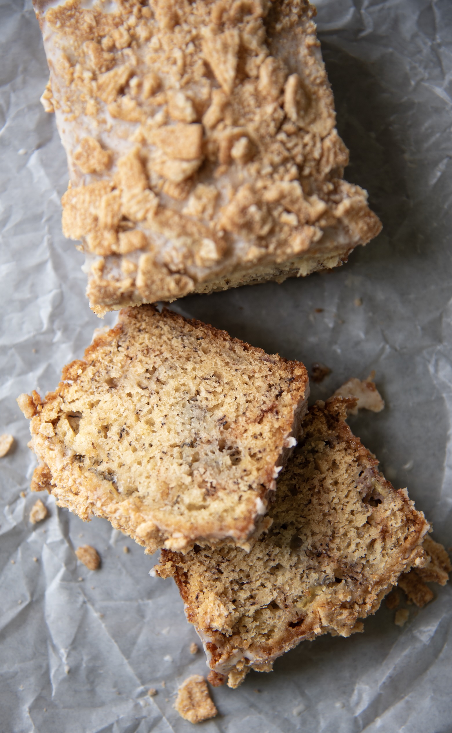 This overhead photo captures a sliced mini loaf of Cinnamon Toast Crunch Banana Bread laid out on crinkled parchment paper. The loaf, topped with a thick layer of crushed Cinnamon Toast Crunch cereal and light glaze, has been cut into several slices, two of which are positioned slightly apart to reveal the bread’s moist, speckled interior. The slices showcase a soft, fluffy crumb flecked with banana and cinnamon, while cereal crumbs and glaze flakes are scattered naturally around them, adding a rustic and inviting touch to the presentation. The overall feel is cozy, homemade, and undeniably delicious.
