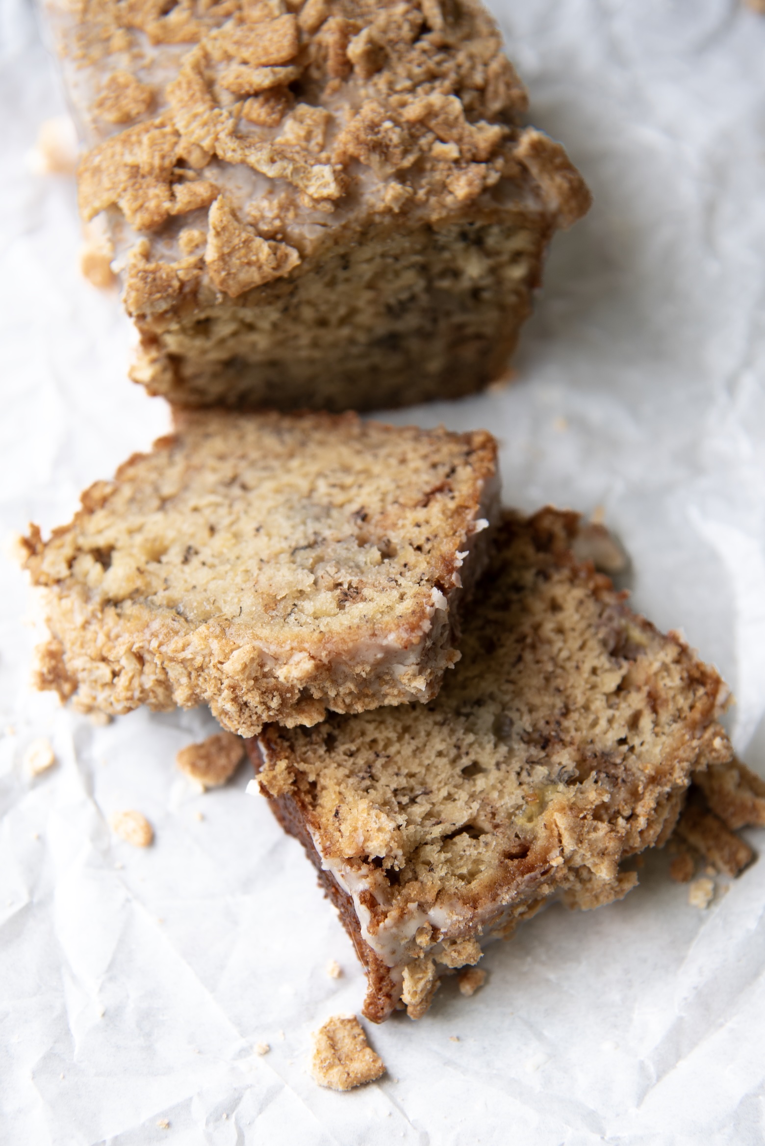 This image features a mini loaf of Cinnamon Toast Crunch Banana Bread on a sheet of crinkled parchment paper, partially sliced to reveal its soft, moist interior. The loaf is topped with a thick coating of crushed Cinnamon Toast Crunch cereal and a light cereal milk glaze that adds texture and sweetness. Two slices have been cut and placed slightly askew in front of the loaf, showing the tender, banana-speckled crumb and lightly browned edges. Scattered cereal bits and crumbs surround the loaf, enhancing its rustic, homemade charm. The photo captures the perfect balance of crunch and softness, inviting you to take a bite.