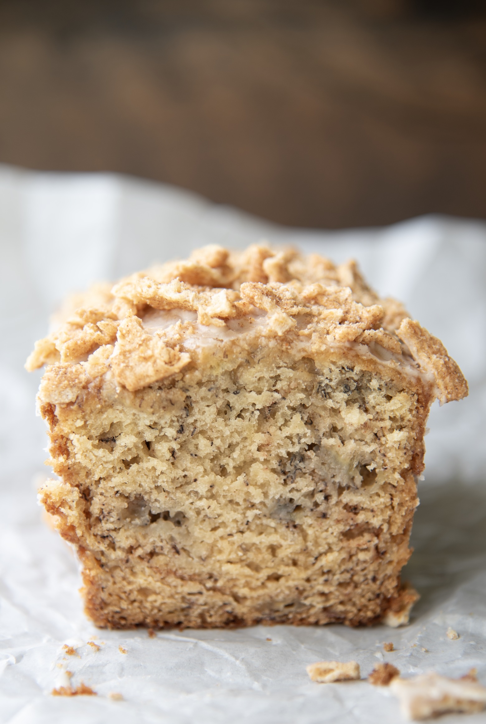 This photo shows a cross-section of a mini loaf of Cinnamon Toast Crunch Banana Bread, set atop a sheet of crinkled parchment paper. The inside of the loaf reveals a moist, tender crumb speckled with banana and perfectly baked to a golden hue. The top of the bread is crowned with a visible layer of crushed Cinnamon Toast Crunch cereal, lightly glazed and adding a crunchy texture that contrasts beautifully with the soft interior. A few crumbs scattered around the base add a casual, homemade touch to the composition. The overall image captures the inviting, comforting essence of freshly baked banana bread with a playful cereal twist.