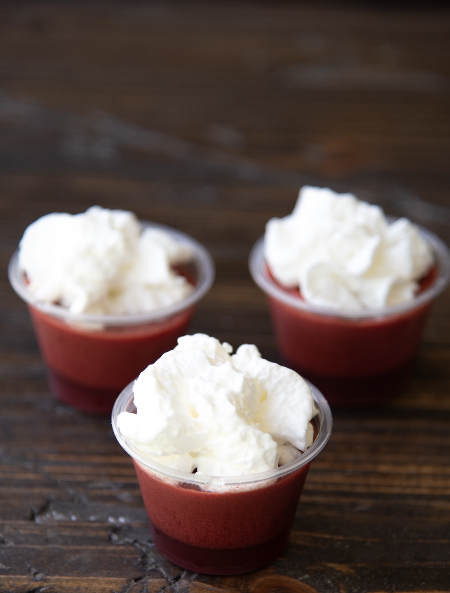 Three small plastic cups filled with layered red jello are arranged on a dark wooden surface. Each jello shot is topped with a fluffy swirl of white whipped cream. The cups are positioned in a slightly staggered triangular formation, with the closest one in sharp focus and the others softly blurred in the background, creating a sense of depth.