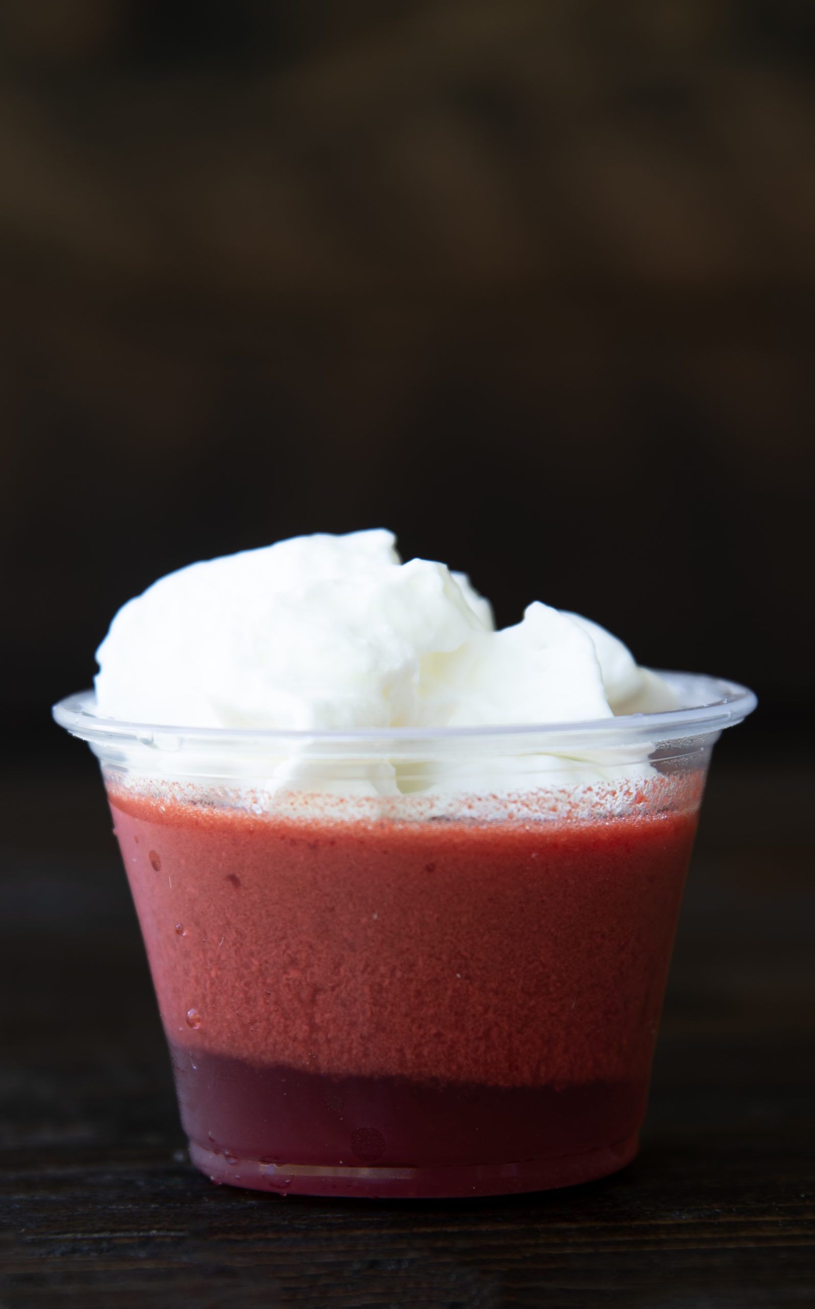 A close-up of a single red layered jello shot in a clear plastic cup, topped with a generous swirl of whipped cream. The jello has a darker red layer on the bottom and a lighter, slightly frothy red layer on top. The background is dark and blurred, emphasizing the vivid colors and textures of the dessert.