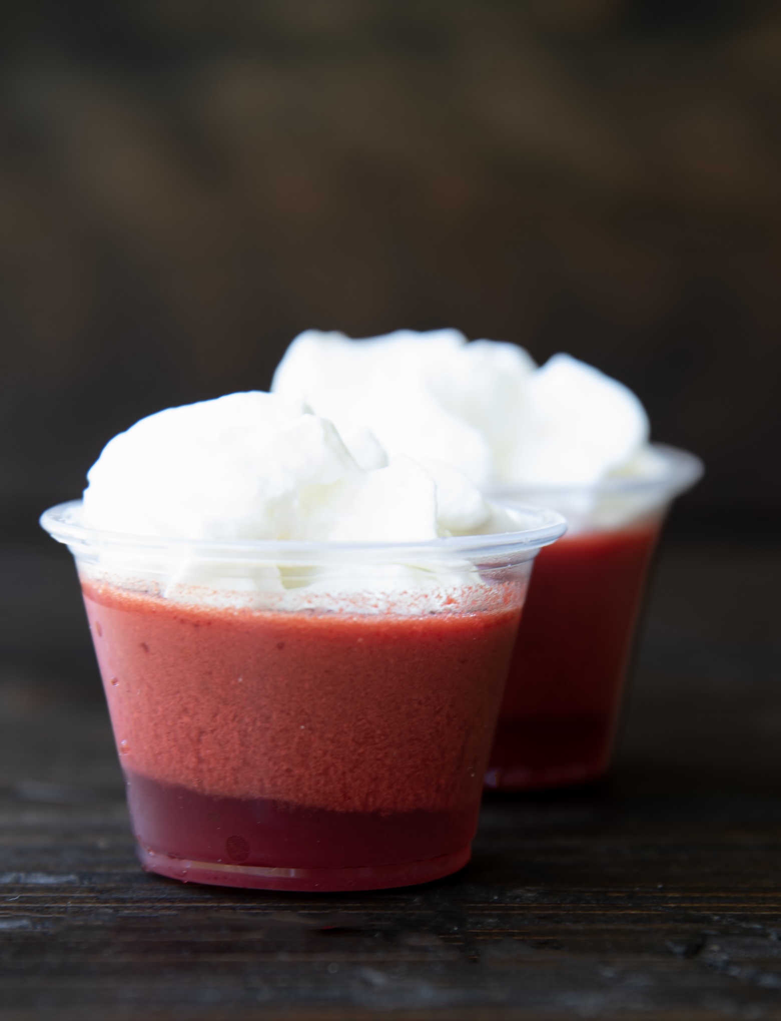 Two red jello shots in clear plastic cups sit on a dark wooden surface. Each dessert features a darker red bottom layer and a lighter, frothy red middle layer, topped generously with fluffy white whipped cream. The front cup is in sharp focus, while the second cup behind it is slightly blurred, creating depth in the composition. The dark background enhances the vibrant colors of the jello and cream.