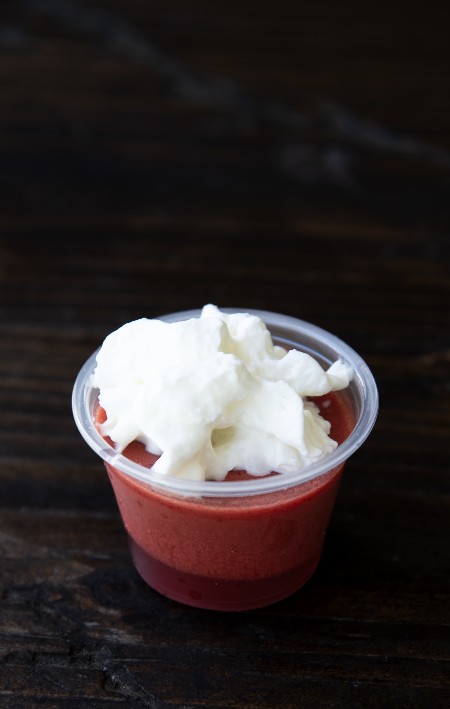 A single red jello shot is displayed in a small clear plastic cup, topped with a fluffy swirl of white whipped cream. The dessert has two distinct red layers—a darker, firmer bottom and a lighter, airy top layer—creating a visually appealing contrast. It rests on a dark wooden surface with a rustic texture.