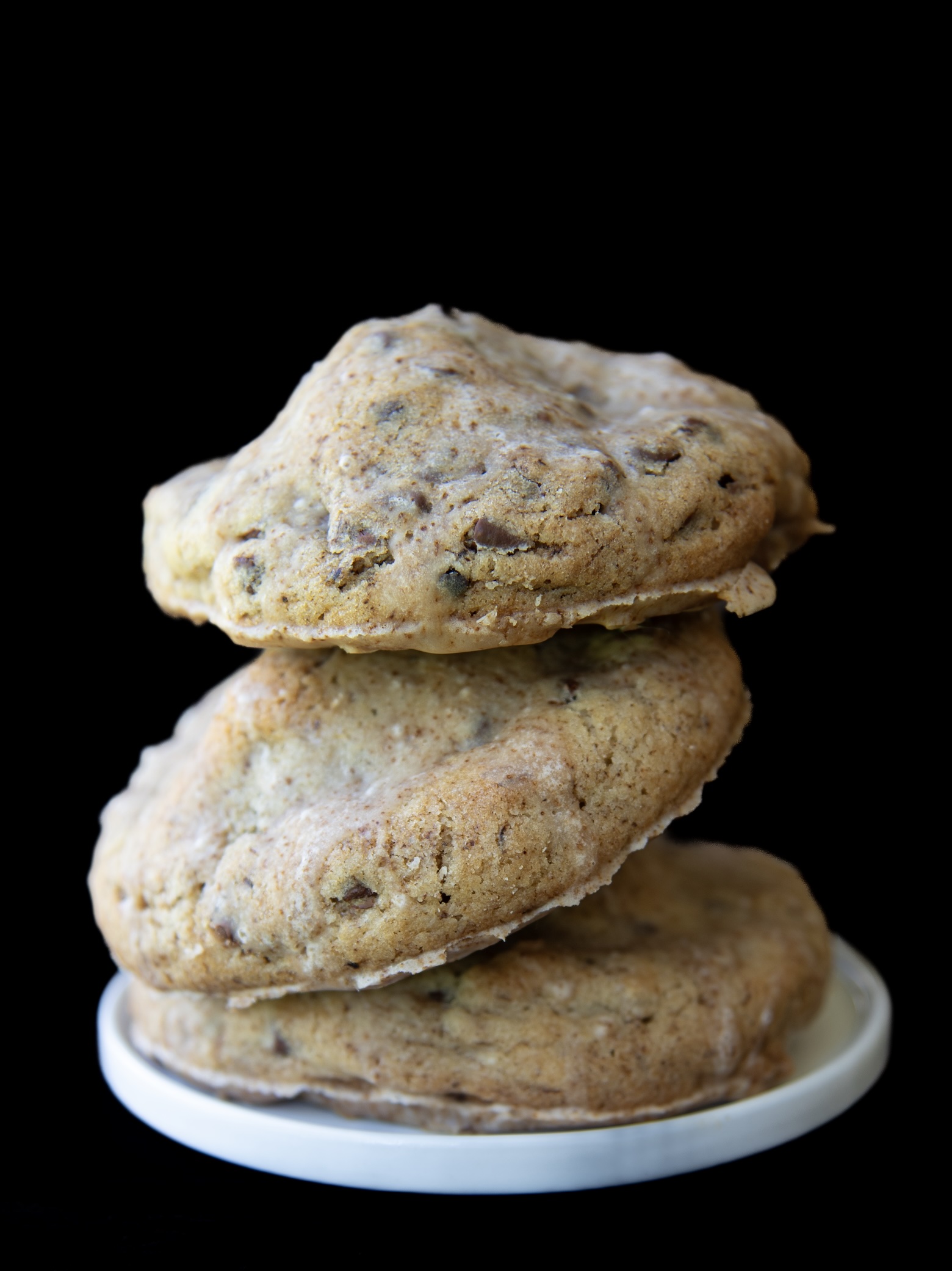 A stack of three large, thick cookies with a golden-brown color, studded with chocolate chips and lightly glazed with a shiny coating. The cookies are irregularly shaped and appear soft and slightly gooey inside. They are balanced on a small white dish against a solid black background, emphasizing their texture and size.