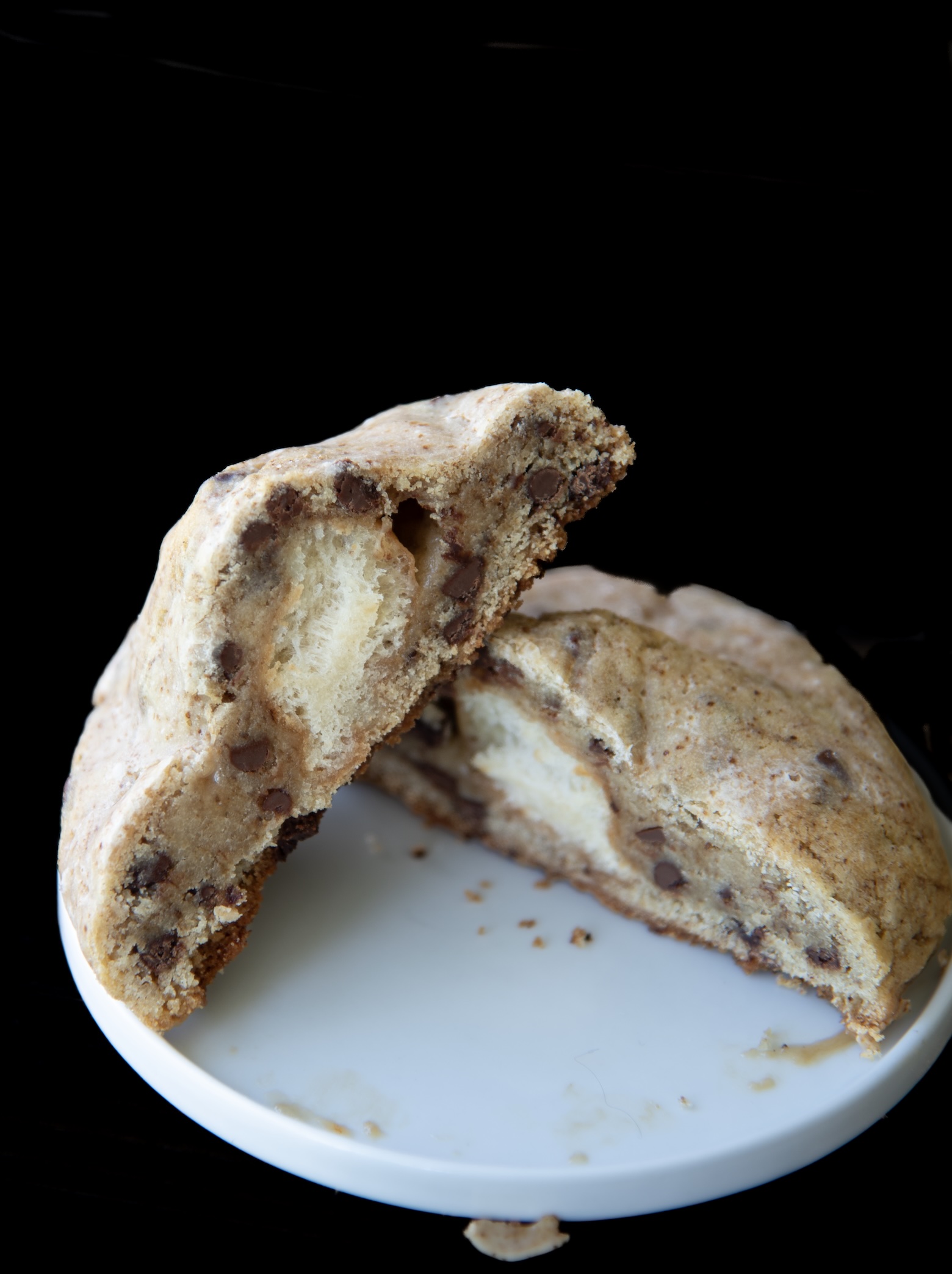 A thick chocolate chip cookie sliced in half and stacked on a small white plate, revealing a soft, fluffy donut-like center surrounded by dense, golden-brown cookie dough. The cookie is generously studded with melted chocolate chips, and a few crumbs are scattered on the plate. The background is completely black, emphasizing the cookie’s texture and indulgent donut filling.