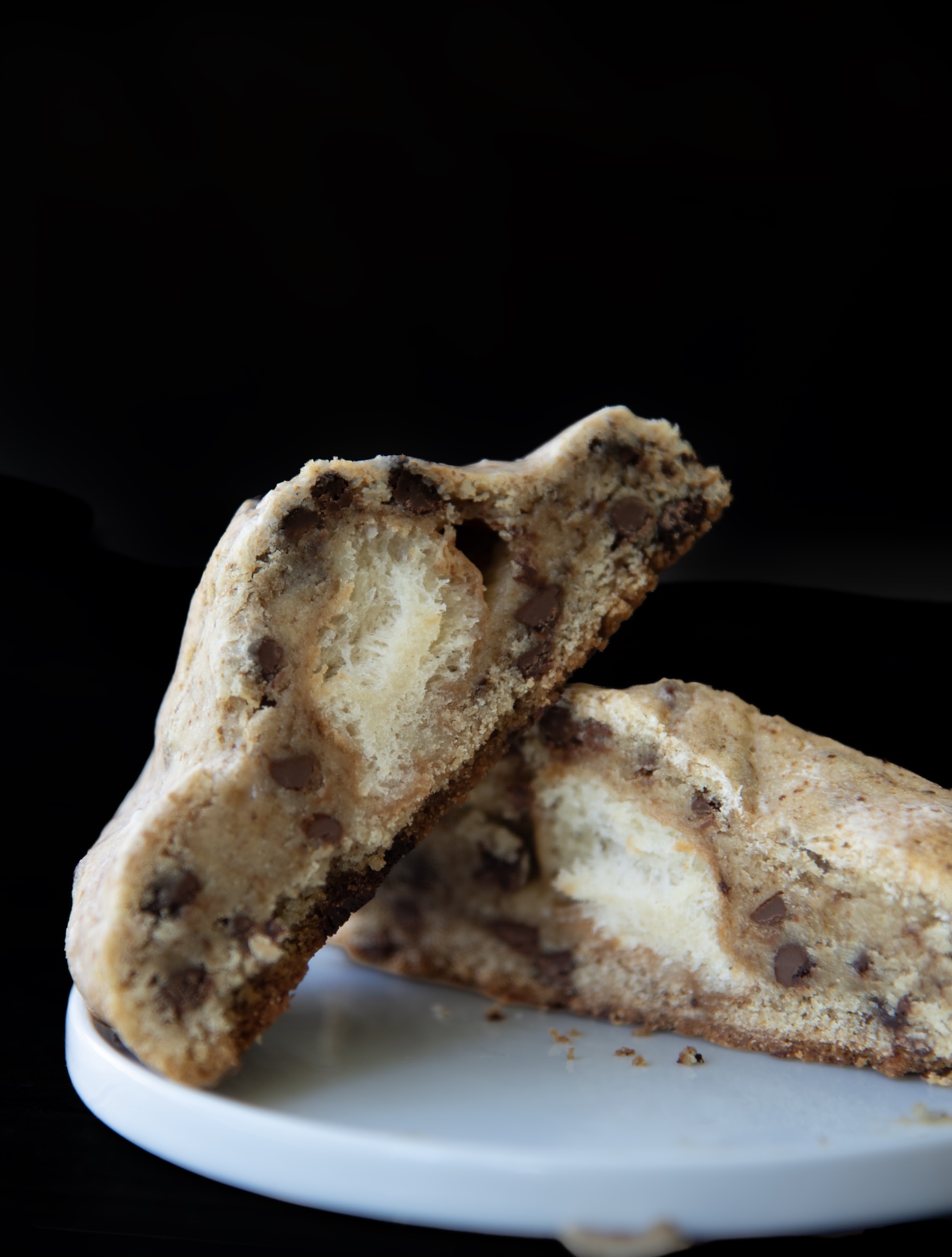 A close-up view of a large cookie split in half, revealing a soft, fluffy donut-like center surrounded by thick, chocolate chip-studded cookie dough. The texture contrast between the airy inner layer and the dense outer cookie is clearly visible. The halves are stacked on a small white plate against a dark background, highlighting the decadent and unique stuffed design.