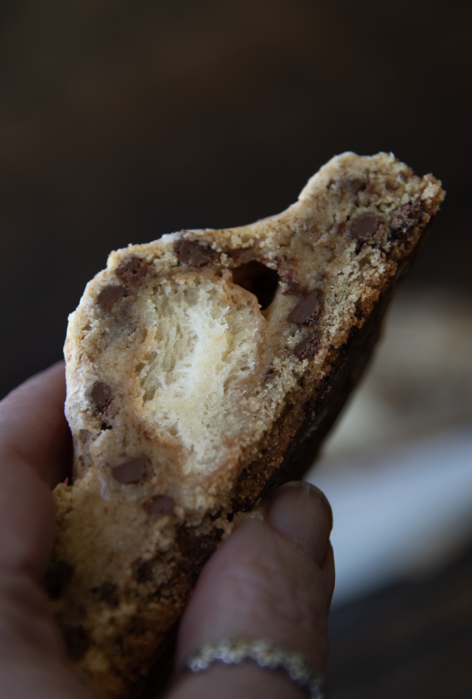 A close-up image of a hand holding half of a thick chocolate chip cookie, revealing a soft, airy donut-like center. The cookie is golden-brown with visible chocolate chips embedded in the dough. The fluffy inner layer contrasts with the chewy outer texture. The background is softly blurred, keeping focus on the detailed texture of the cookie and the hand holding it, which wears a delicate silver ring.