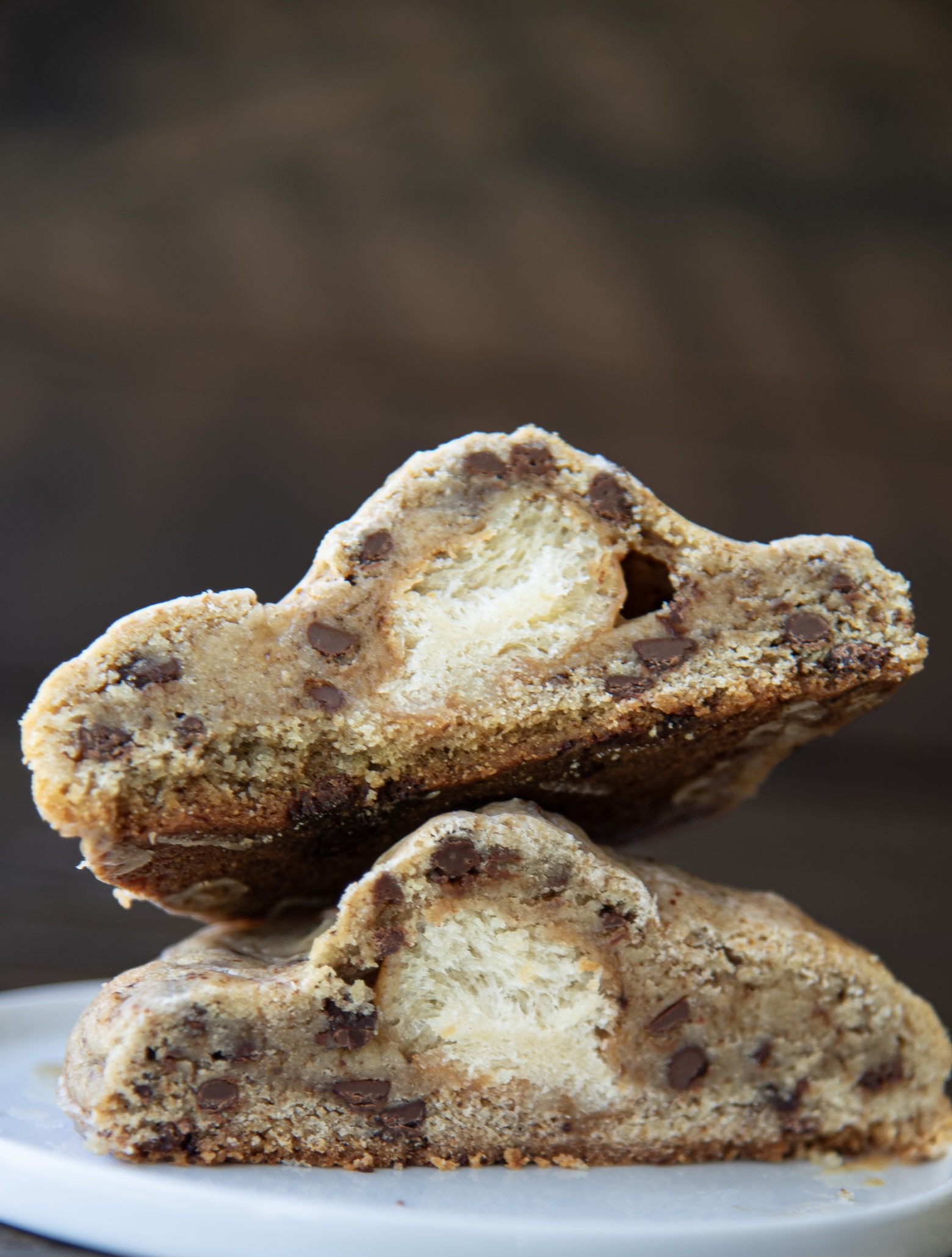 A close-up of a cookie sliced in half and stacked, showcasing a unique filling of fluffy donut bread inside a thick, golden-brown chocolate chip cookie. The cookie exterior is dense and studded with chocolate chips, while the center reveals a soft, airy interior with a distinct texture contrast. The image is set against a softly blurred brown background, with the halves resting on a smooth white plate.