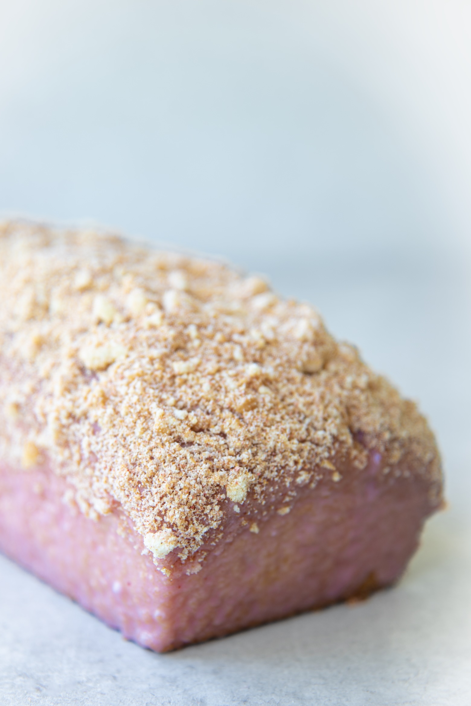 Close up side view of a whole strawberry shortcake loaf cake sitting on a light gray surface. The cake is covered in a pink strawberry glaze and generously topped with a golden crumb coating made of crushed cookies and freeze-dried strawberries. The texture of the glaze and crumbs is clearly visible.