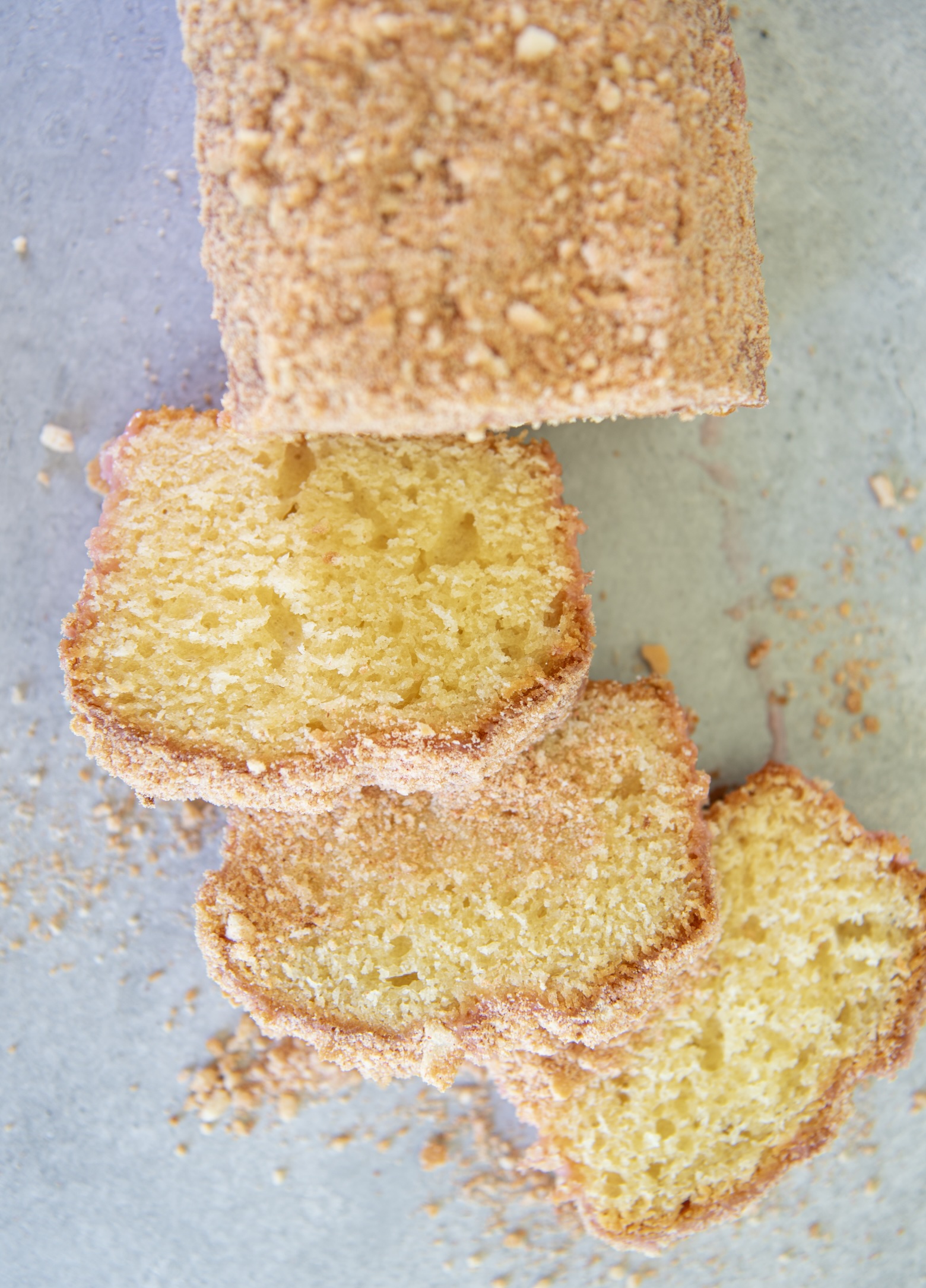 Overhead view of a sliced strawberry shortcake loaf cake on a light gray surface. The slices reveal a moist, fluffy yellow interior with a pink glaze and a golden cookie-strawberry crumb topping. Crumbs are scattered around the cake, emphasizing its crunchy texture.