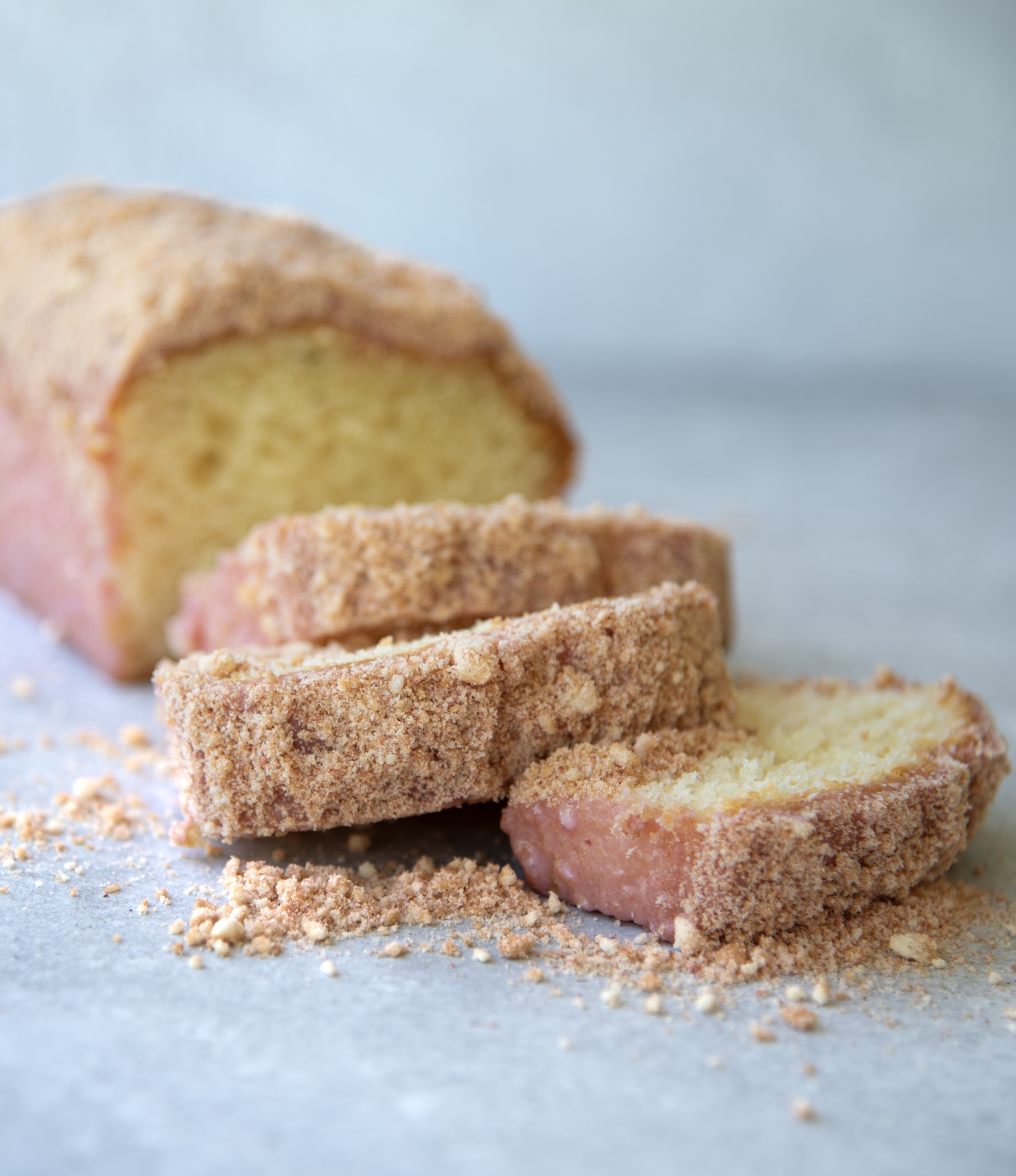Close-up of a sliced strawberry shortcake loaf cake on a light surface. The cake has a pink strawberry glaze and is coated with a golden strawberry-cookie crumb topping. Crumbs are scattered around the slices, and the texture of the moist yellow cake interior is visible.