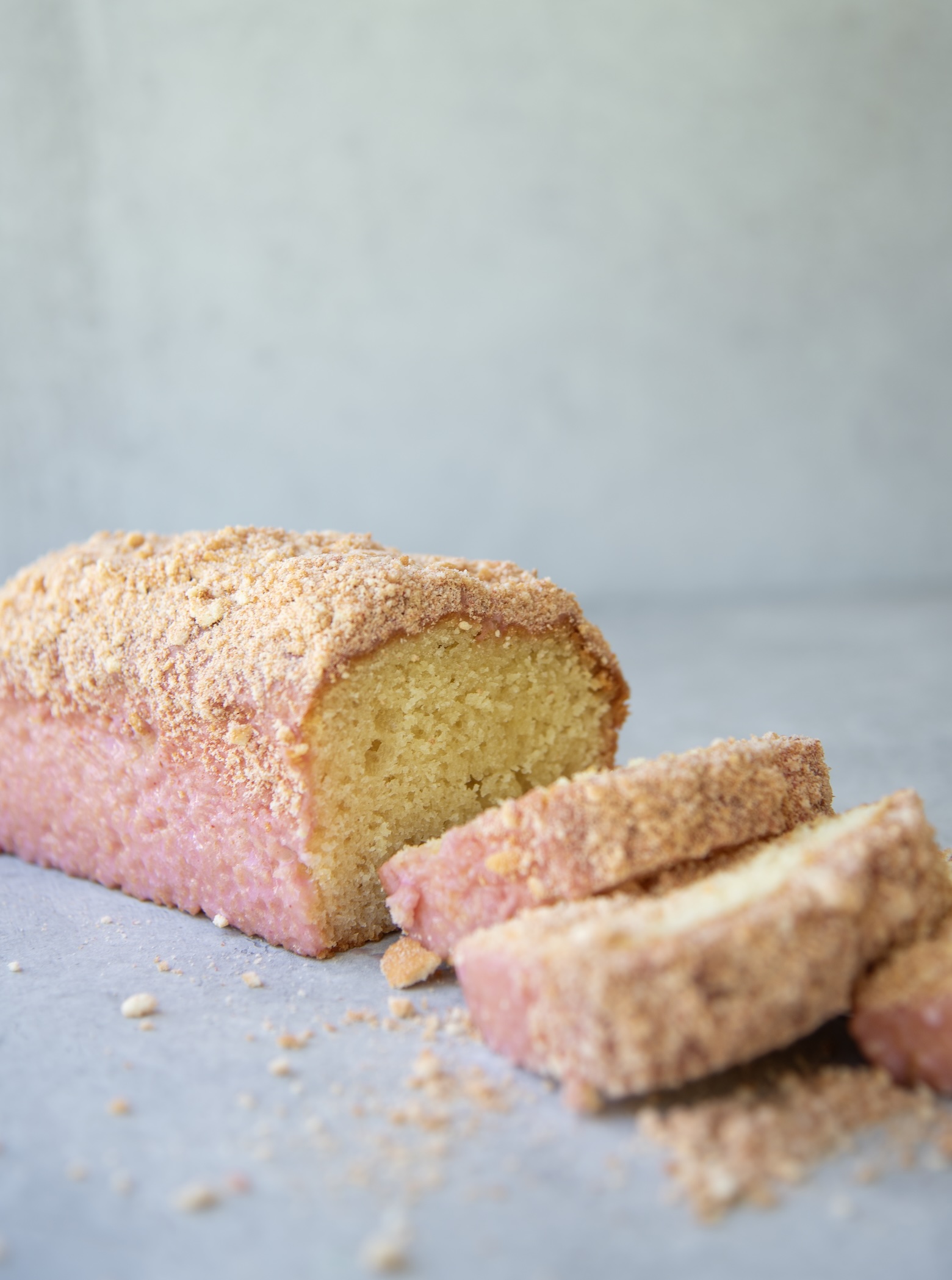 A whole strawberry shortcake loaf cake with a few slices cut and fanned out in the foreground. The loaf is coated in a pink strawberry glaze and covered with a golden crumb topping. Crumbs are scattered around the base on a light gray surface, and the interior of the cake appears moist and fluffy.
