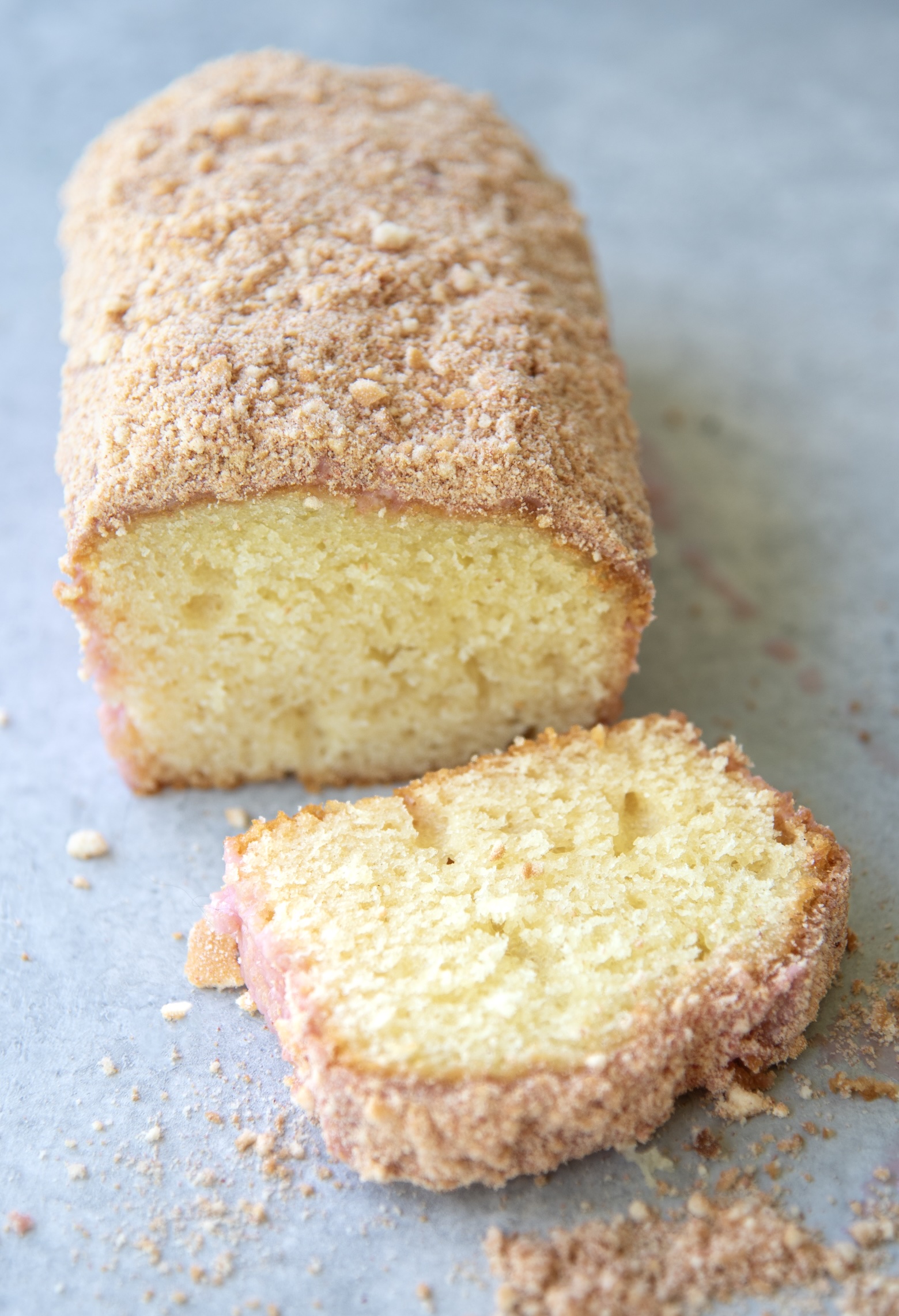 Strawberry shortcake loaf cake with one slice cut and resting in front. The cake has a soft yellow interior, a light pink glaze around the edges, and is topped with a golden strawberry-cookie crumb. Crumbs are scattered across the light gray surface.
