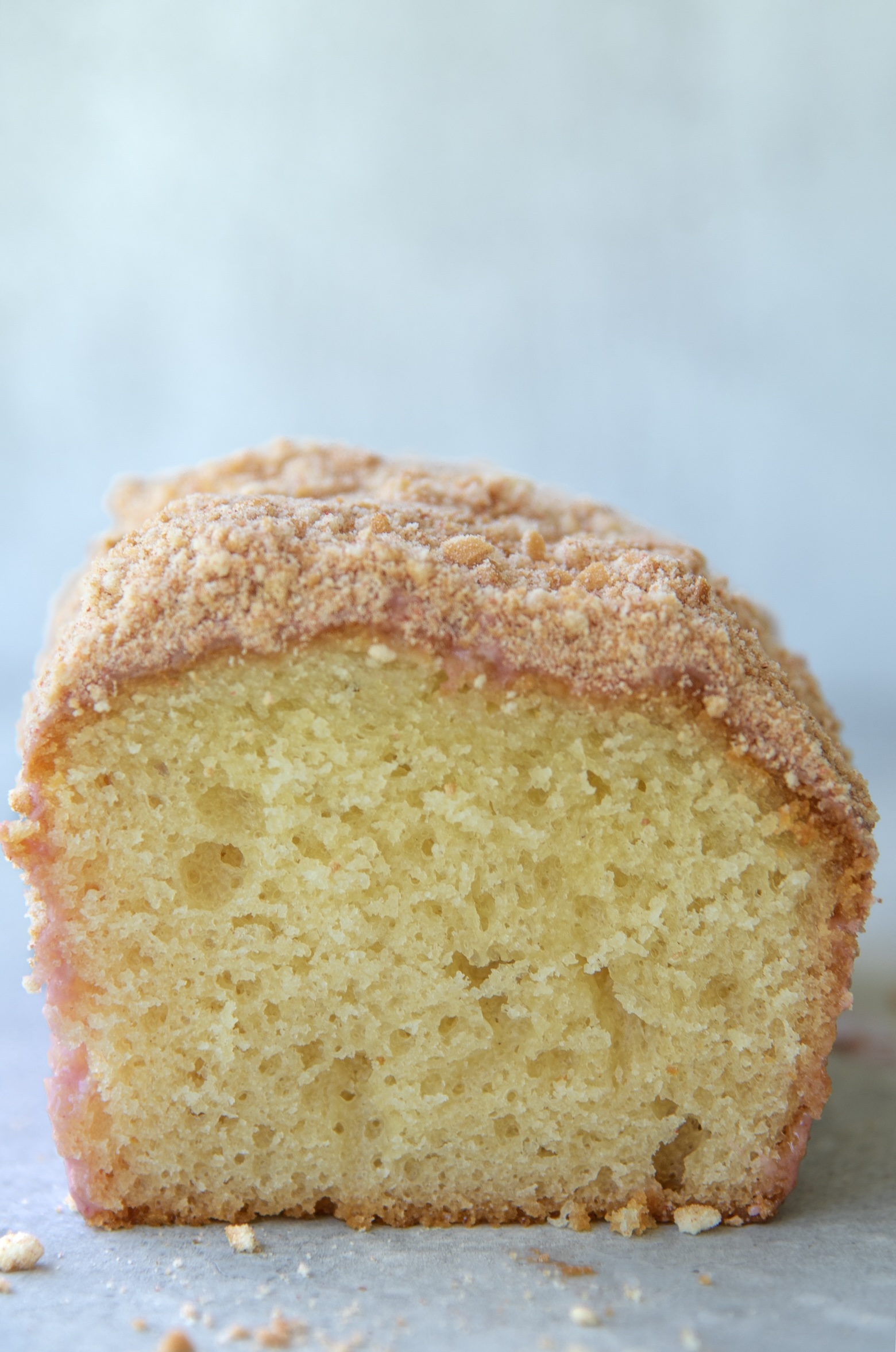 Close up view of the front of a strawberry shortcake loaf cake, showing its moist, golden yellow interior. The cake is topped with a pink glaze and a crumbly golden cookie strawberry crumb layer. A few crumbs are scattered on the light gray surface beneath the cake.