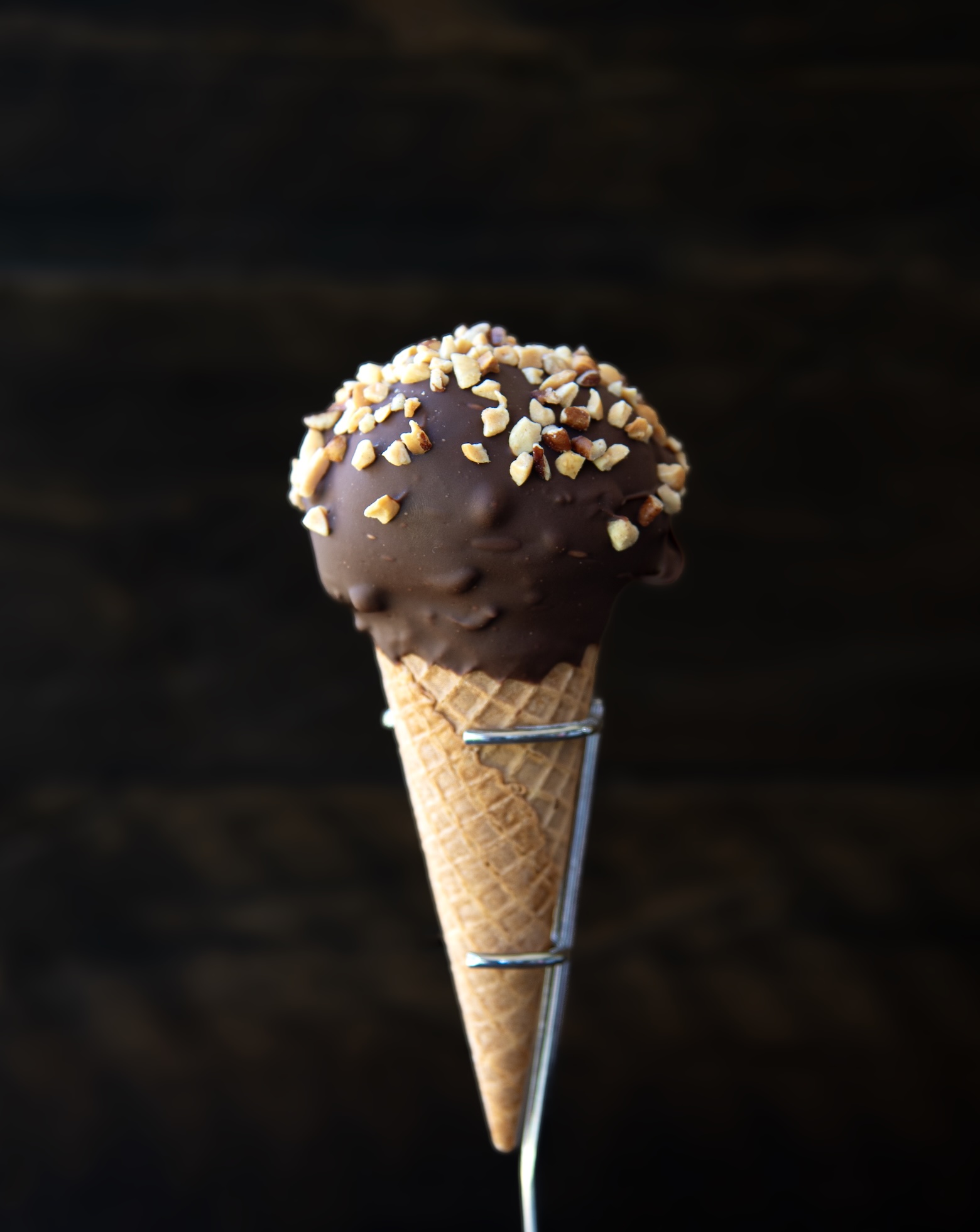 A centered image of an ice cream cone held upright in a metal stand against a dark, softly textured background. The cone features a scoop of vanilla ice cream completely coated in a thick chocolate shell, topped with a generous sprinkle of chopped nuts. The chocolate coating is smooth with a few subtle ridges, giving it a rich, glossy appearance. The waffle cone has a classic grid texture and tapers to a fine point at the bottom, contrasting nicely with the rounded, nut-covered top.
