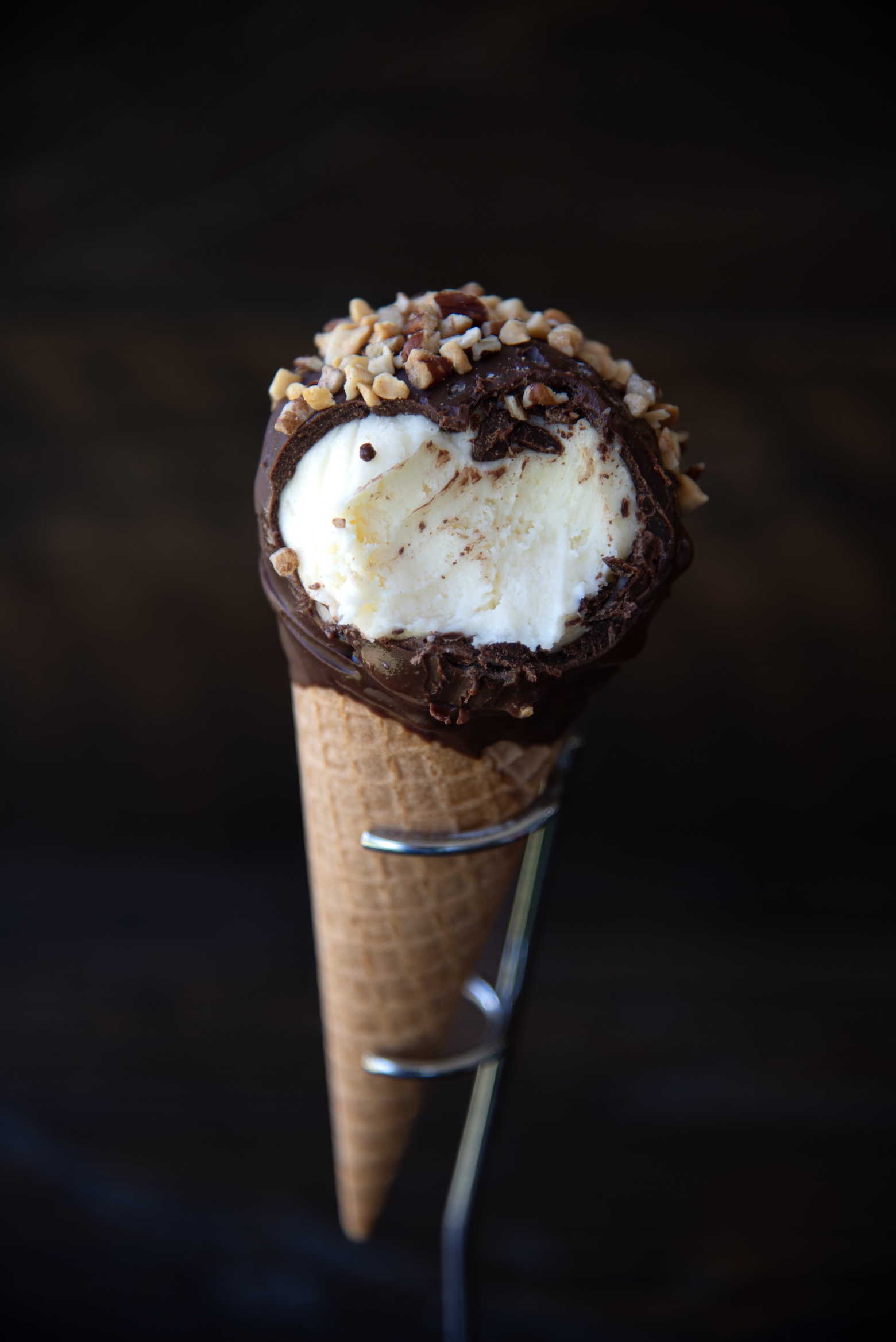 A close-up image of a vanilla ice cream cone held upright in a metal stand against a dark background. The ice cream is coated with a thick layer of chocolate around the top edge, sprinkled generously with chopped peanuts. A bite has been taken out of the ice cream, exposing its creamy interior. The waffle cone has a classic grid texture and is firmly nestled in the metal holder, making the dessert the central focus of the composition.