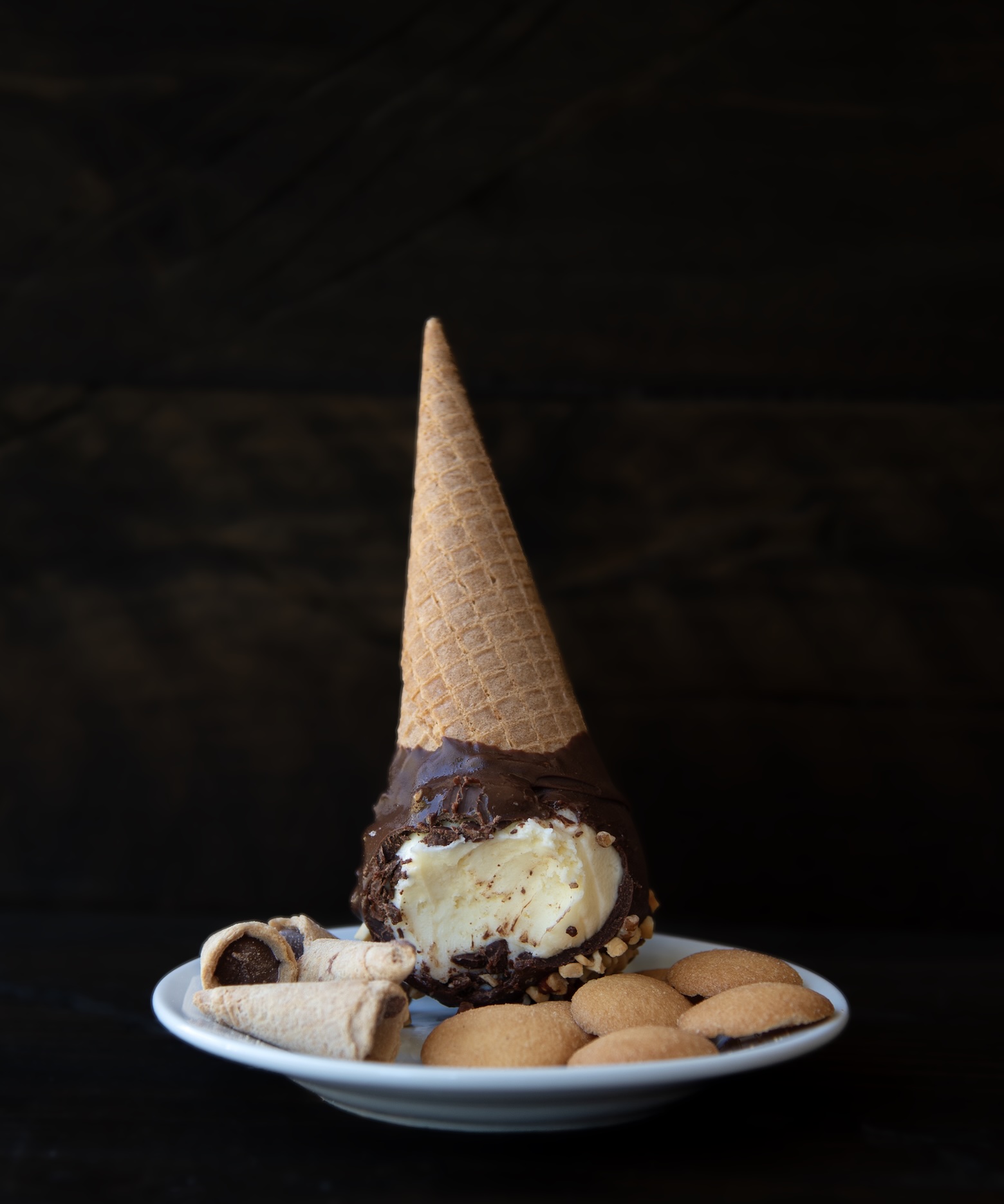 A whimsical dessert scene featuring an ice cream cone placed upside down on a white plate. The cone is filled with vanilla ice cream, partially dipped in chocolate and sprinkled with chopped nuts, with a bite taken out of the ice cream. Surrounding the cone are pieces of rolled wafer cookies filled with chocolate and several round vanilla wafer cookies. The dark, moody background contrasts with the light colors of the dessert, making it the focal point of the image.