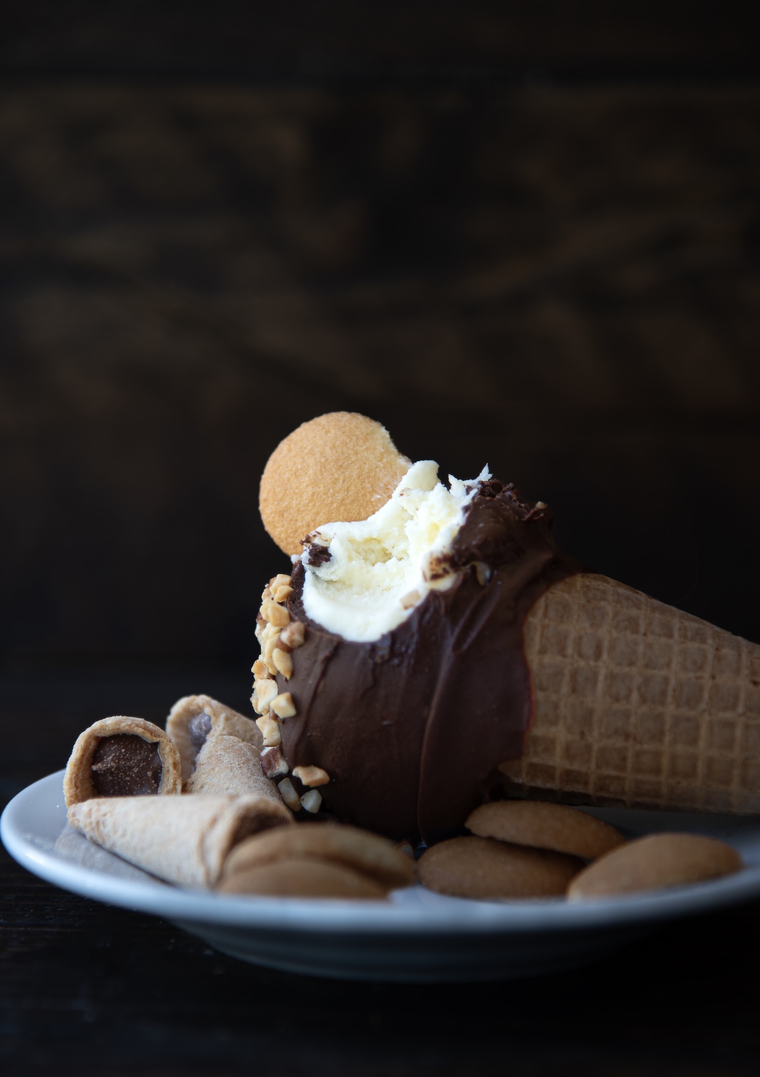 A close-up image of an ice cream cone dessert resting on a white plate. The cone is filled with vanilla ice cream and topped with a thick chocolate coating, chopped peanuts, and a vanilla wafer cookie. A bite has been taken from the ice cream, revealing its creamy interior. Surrounding the cone on the plate are broken pieces of waffle cone and several whole vanilla wafer cookies.