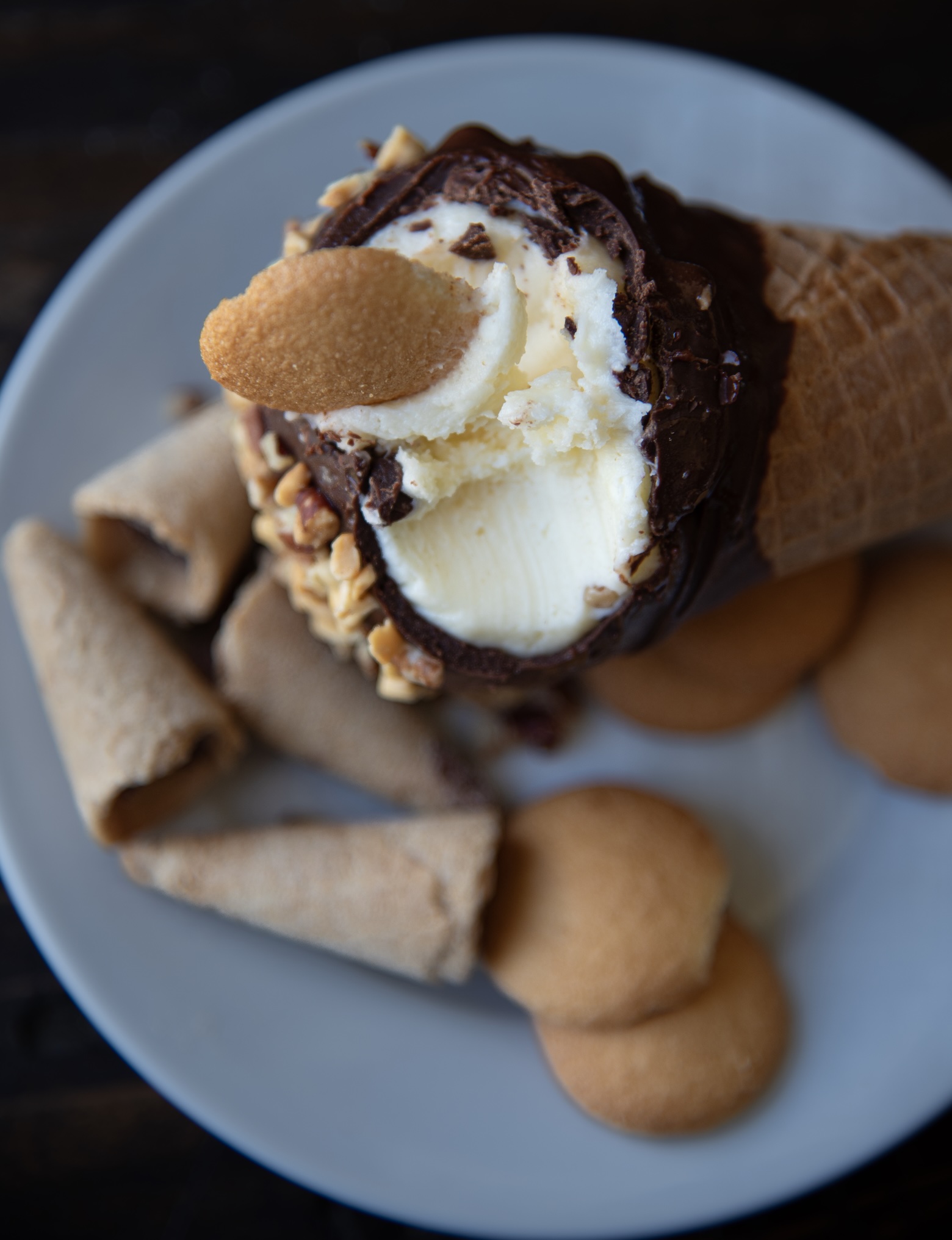 A close-up photo of a deconstructed ice cream cone dessert on a white plate. The focal point is a vanilla ice cream cone with a bite taken out, topped with a chocolate coating, chopped nuts, and a vanilla wafer cookie. Surrounding the cone are additional broken waffle cone pieces and more vanilla wafer cookies, arranged casually on the plate. 