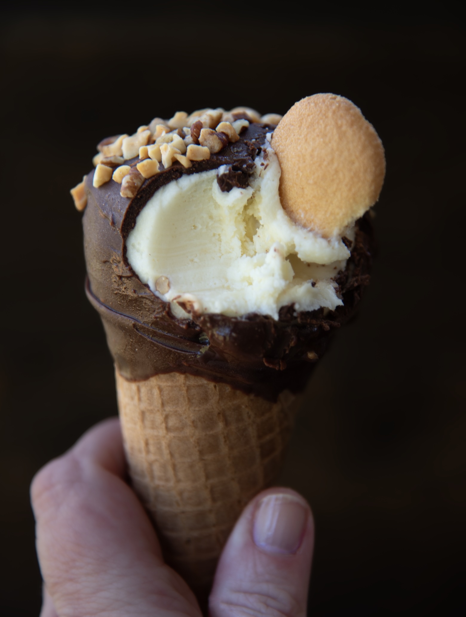 A close-up image of a hand holding an ice cream cone. The cone is topped with vanilla ice cream that has a chocolate coating around the top edge. The chocolate coating is sprinkled with chopped nuts and features a single vanilla wafer cookie pressed into the ice cream. A bite has been taken from the ice cream, revealing its creamy texture. The background is dark and out of focus, emphasizing the dessert.