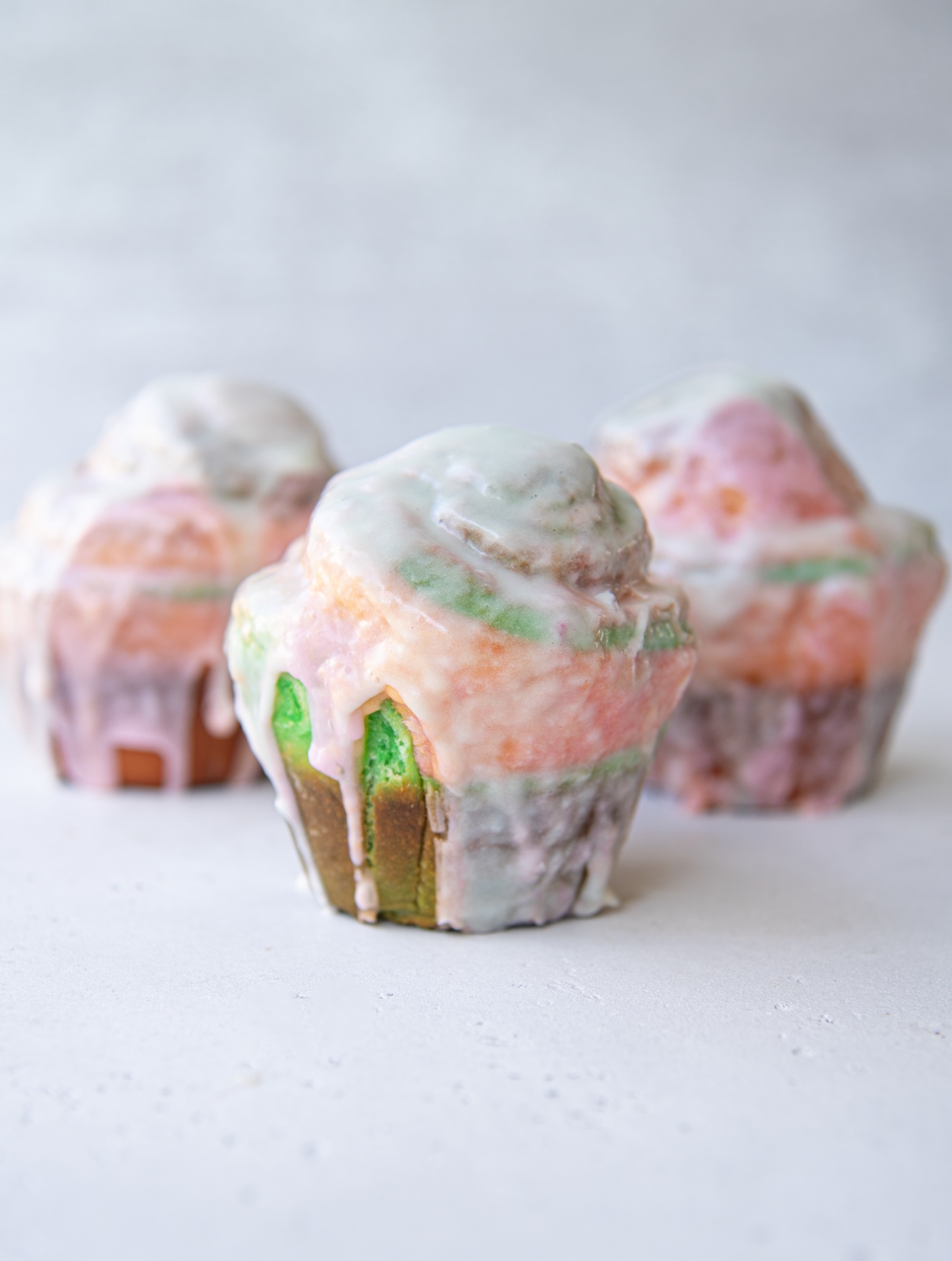 Three rainbow sherbet sweet rolls are displayed on a light surface. The rolls have a swirled, multicolored dough in shades of green, pink, and orange, with a generous coating of white glaze dripping down the sides. The glaze has a glossy finish, and the vibrant colors of the rolls peek through beneath it. The rolls are arranged in a casual cluster, with one prominently in the foreground and two slightly blurred in the background.