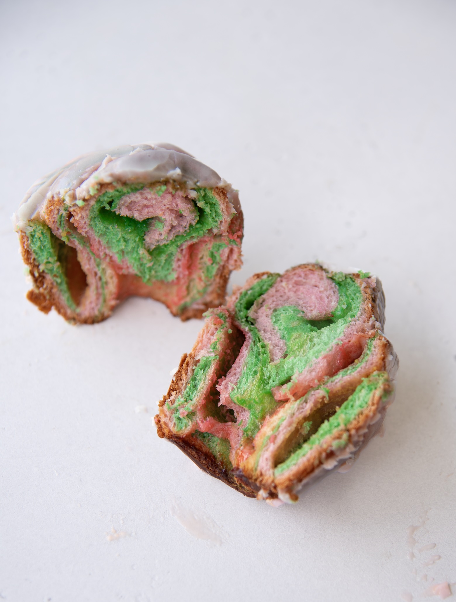 A rainbow sherbet sweet roll is cut in half and placed on a light surface, revealing its vibrant interior. The swirled dough features bright pink and green colors in a marbled pattern, with a soft, fluffy texture. A thin layer of white glaze coats the outer surface of the roll, slightly dripping along the edges. The cross-section showcases the intricate spiral layers of the sweet, colorful pastry.