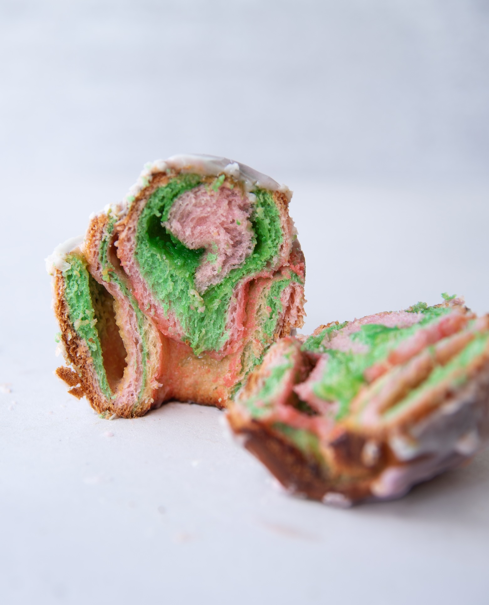 A close-up image of a rainbow sherbet sweet roll sliced in half, revealing vibrant swirls of pink and green dough inside. The interior has a soft, fluffy texture with defined spiral layers, while the outer edge is lightly golden brown. A thin layer of white glaze coats the top, slightly melting over the edges. One half stands upright, displaying its colorful cross-section, while the other lies slightly blurred in the foreground. The background is a clean, light surface, emphasizing the bright colors of the roll.