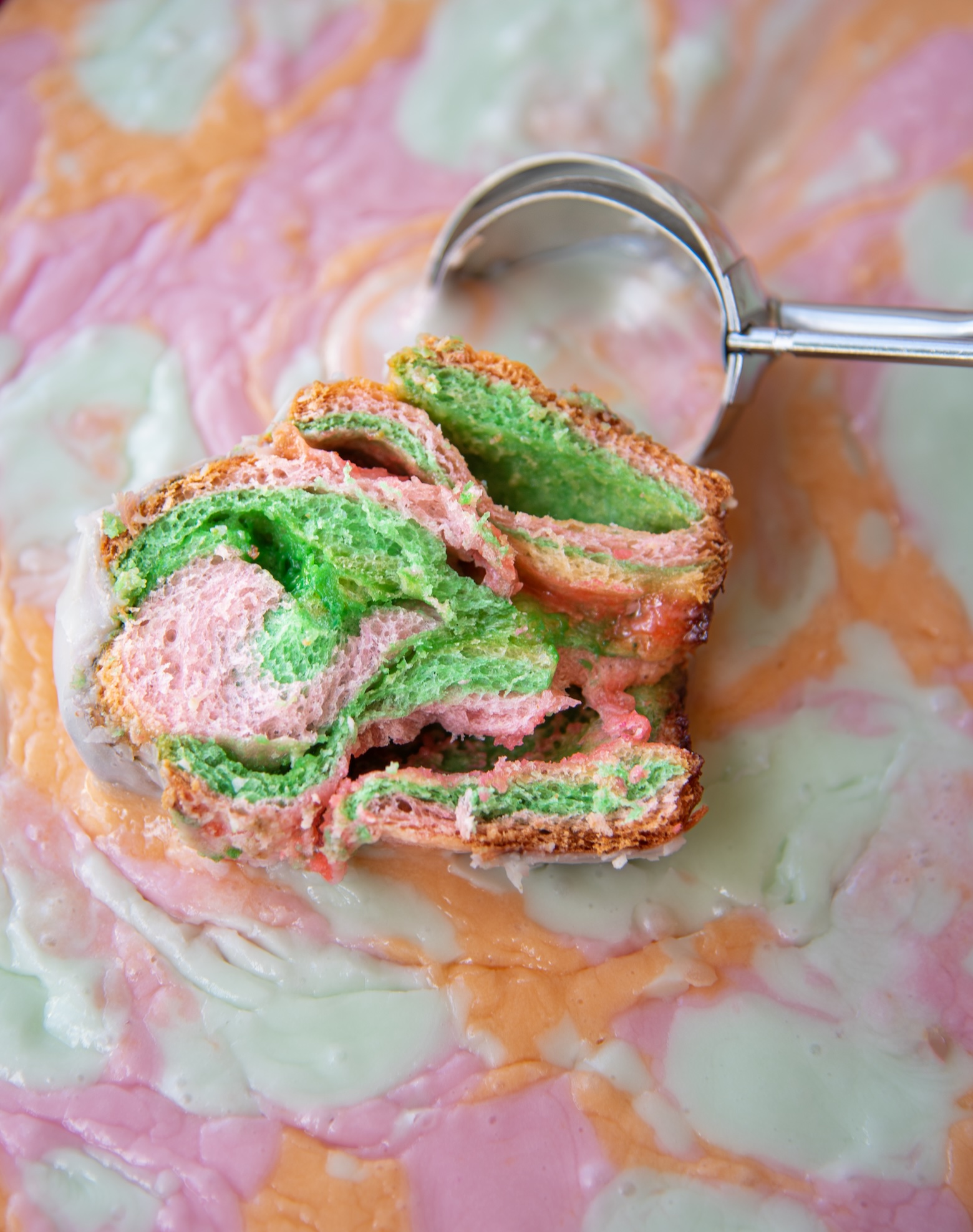 A vibrant slice of a rainbow sherbet sweet roll rests on a colorful, swirled background that resembles melted rainbow sherbet ice cream in shades of pink, orange, and mint green. The roll features vivid pink and green layers of fluffy dough with a lightly crisp, golden-brown edge and a hint of white glaze. A shiny metal ice cream scoop lies just behind the roll, partially resting on the creamy surface, enhancing the playful, dessert-inspired theme of the composition.