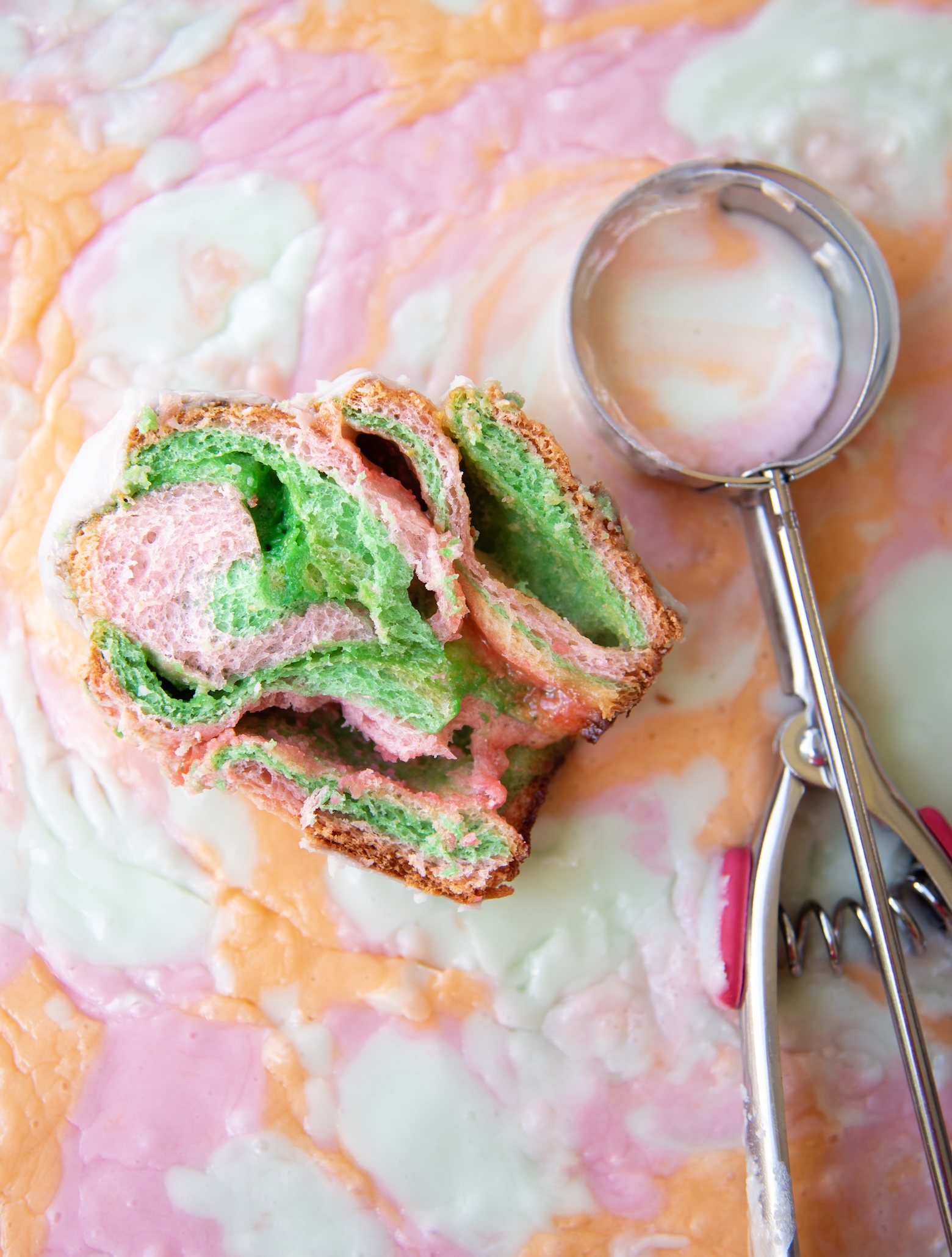 A slice of rainbow sherbet sweet roll is placed on a colorful, swirled surface that resembles melted rainbow sherbet in pastel shades of pink, orange, and mint green. The roll displays soft, airy layers of pink and green dough with a light golden crust and a touch of white glaze on top. Beside it lies a metal ice cream scoop with red handles, partially resting on the creamy, marbled background, adding a playful, dessert-themed touch to the composition.